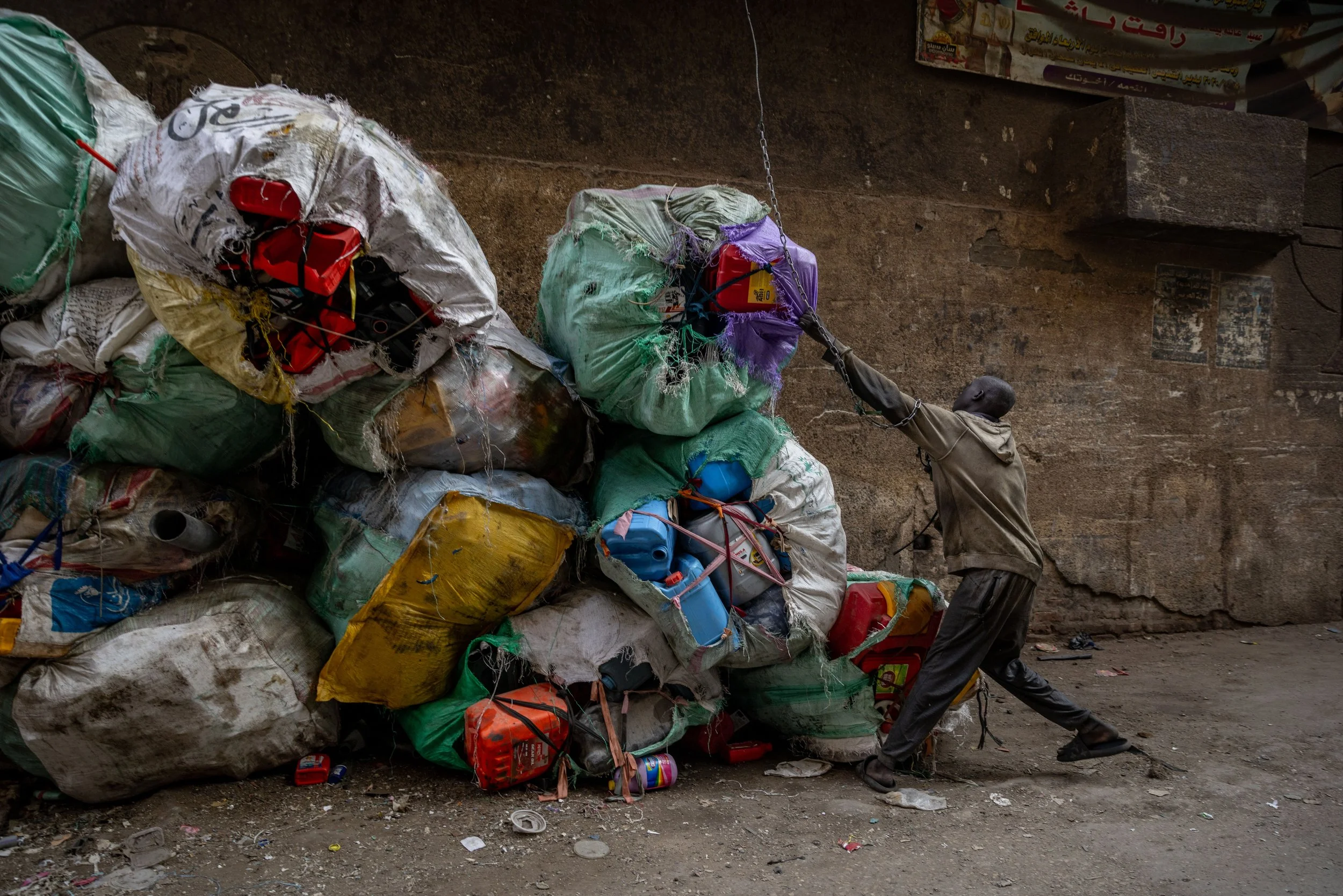 A man in dark clothing is pulling a large rope tied to a pile of colorful, tightly packed garbage bags against a rough, brown wall.
