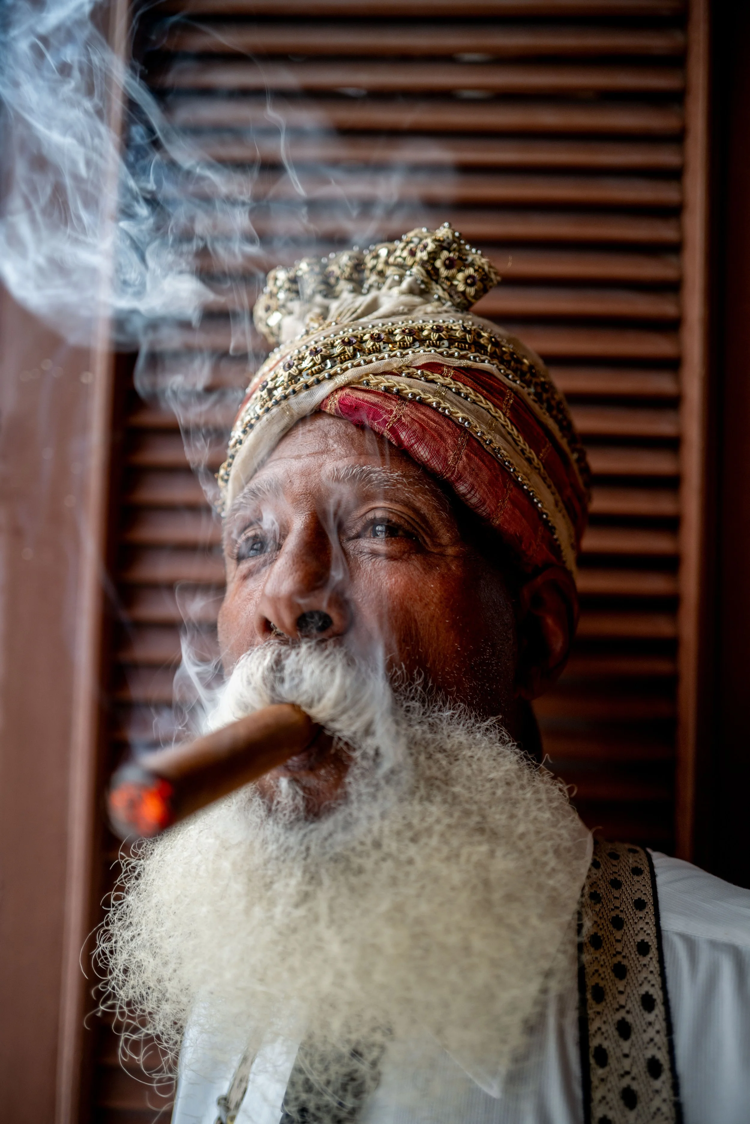 An elderly man with a long white beard and mustache is smoking a cigar. He is wearing a traditional, ornate turban and a white shirt with embroidered detail on the shoulder. Smoke is visible around his face, and he appears to be looking slightly upwa