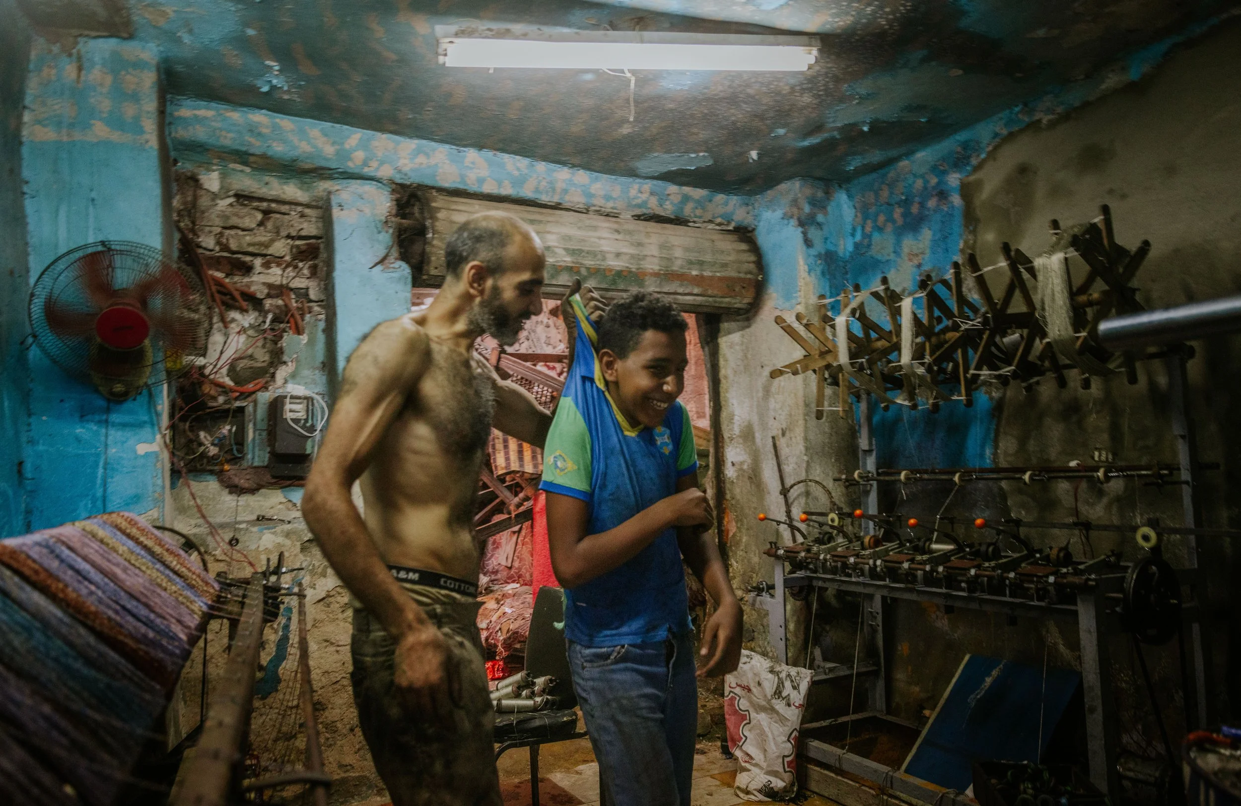 Two men, one shirtless and another wearing a blue and green t-shirt, smiling and interacting inside a small, rustic workshop with worn blue-painted walls, tools on the wall, and machinery in the background.