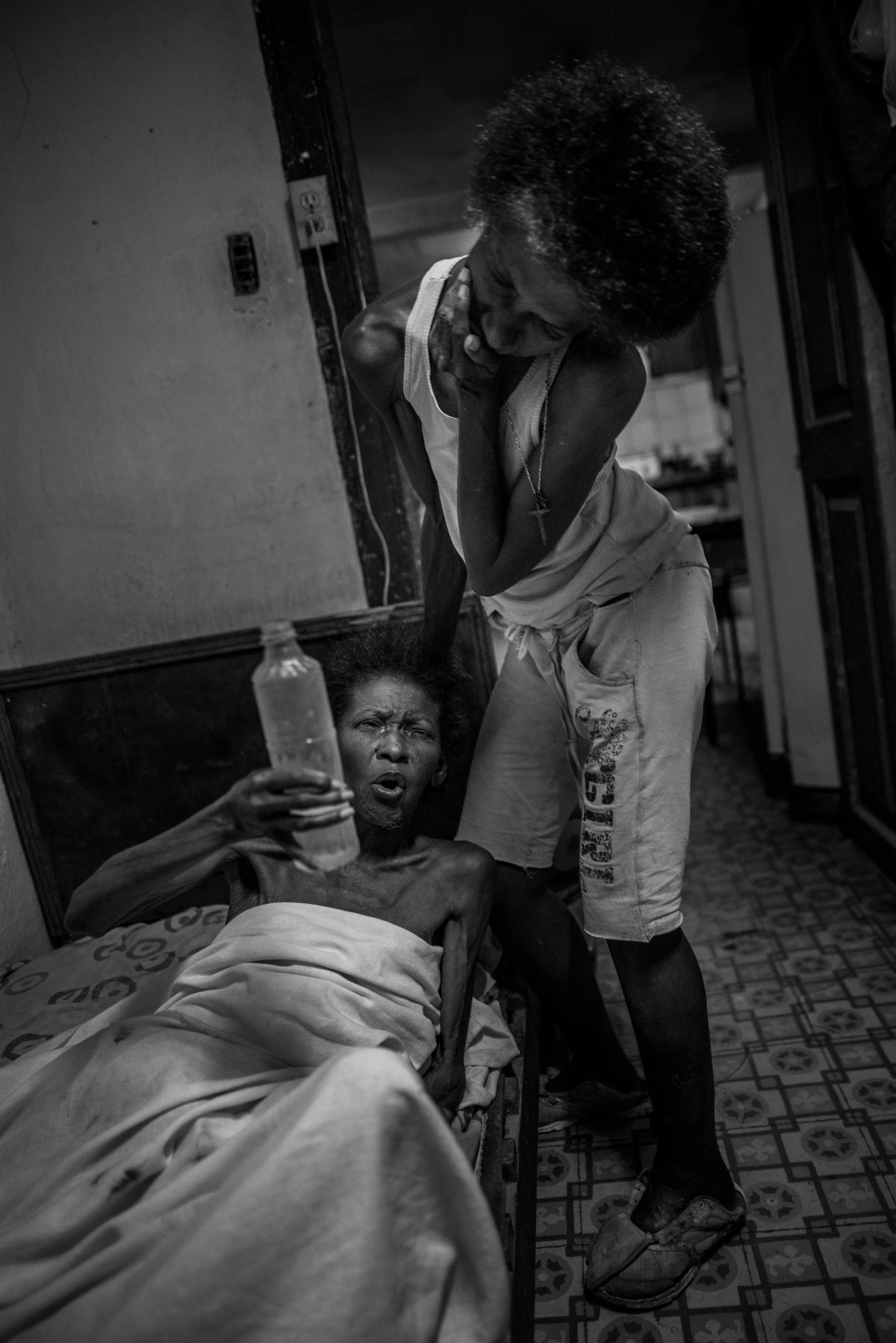 A woman lying in a hospital bed holding a water bottle, with a distressed expression, while a man bending over her appears to be comforting or attending to her.