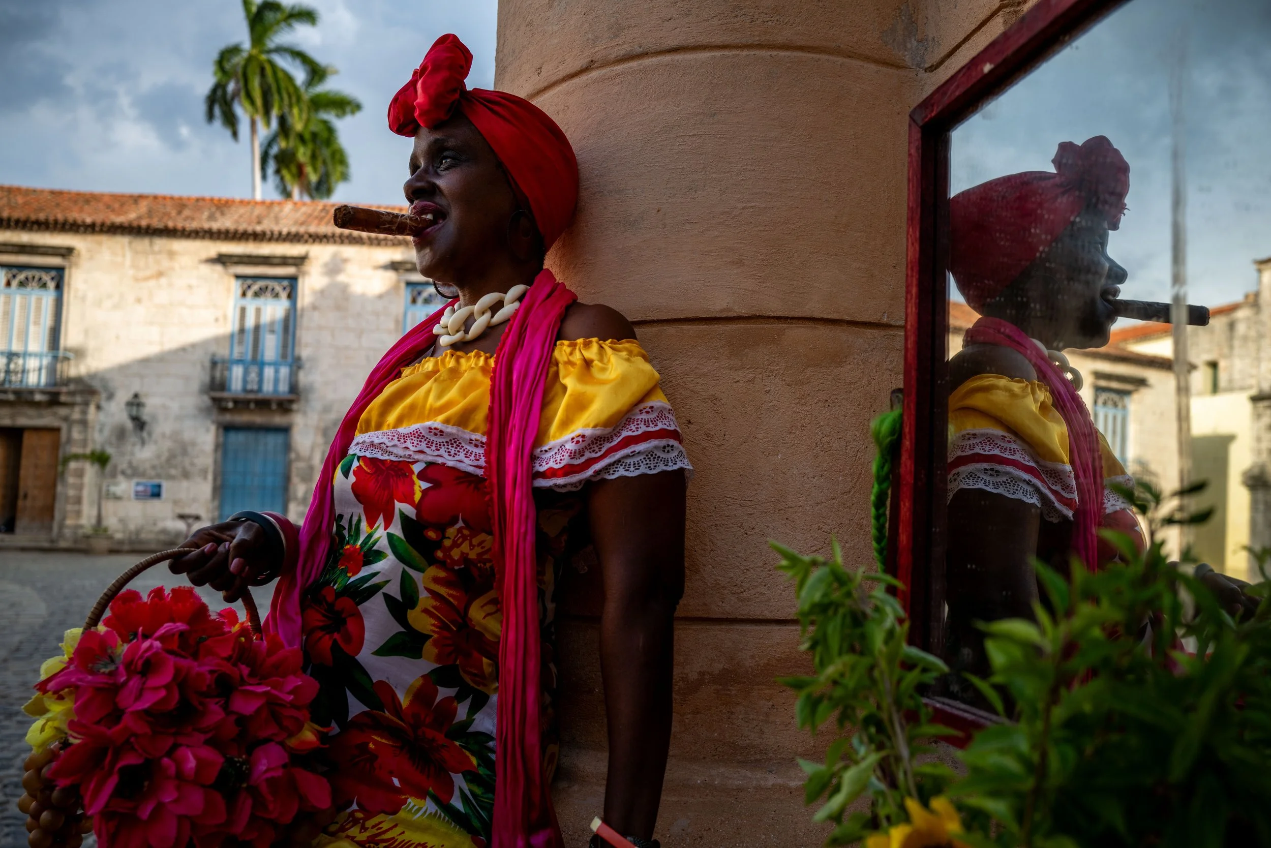 An elderly woman in colorful traditional clothing and jewelry is standing outside a building, holding a basket of pink flowers. She has a cigar in her mouth and is wearing a red headscarf and a yellow top with floral patterns. The woman is leaning ag