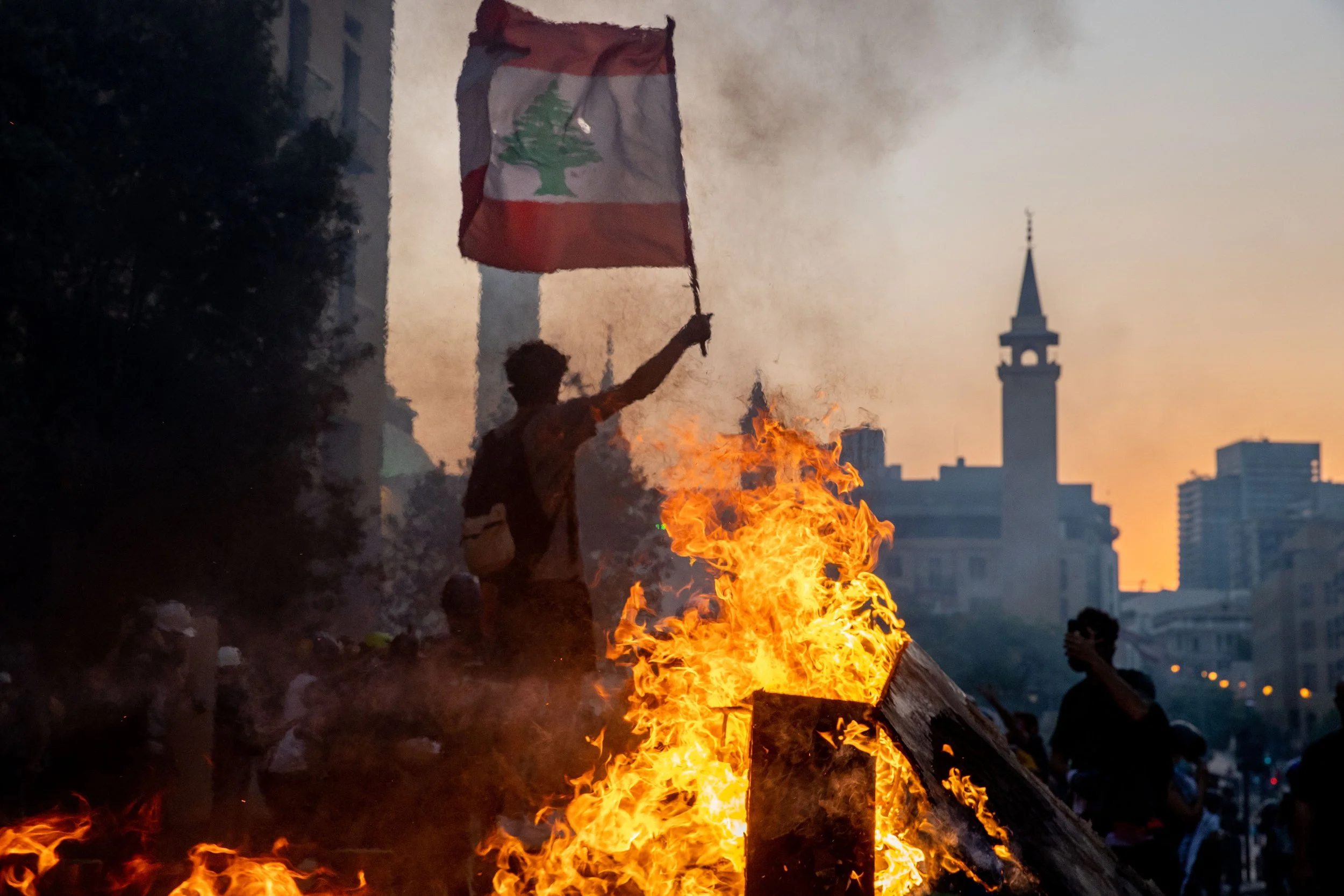 Person holding a Lebanese flag above a fire during a protest or demonstration at sunset, with a city skyline and a prominent clock tower in the background.