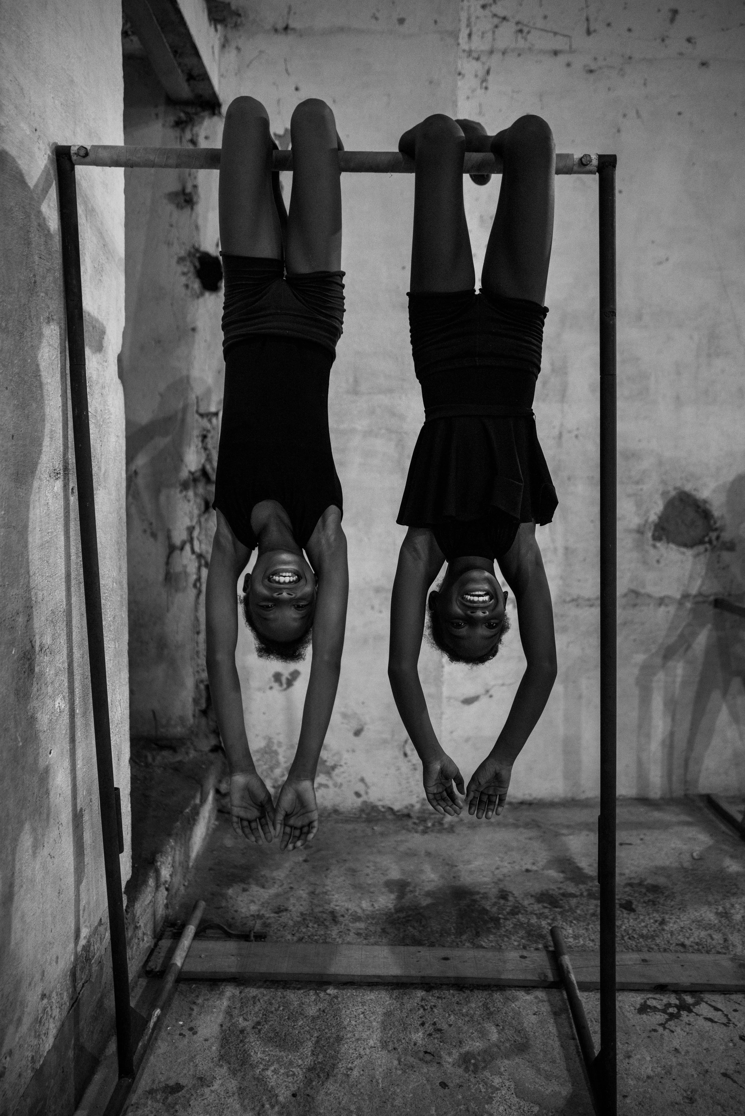 Two young girls hanging upside down on a gym bar, smiling and looking at the camera, in a gritty indoor setting.