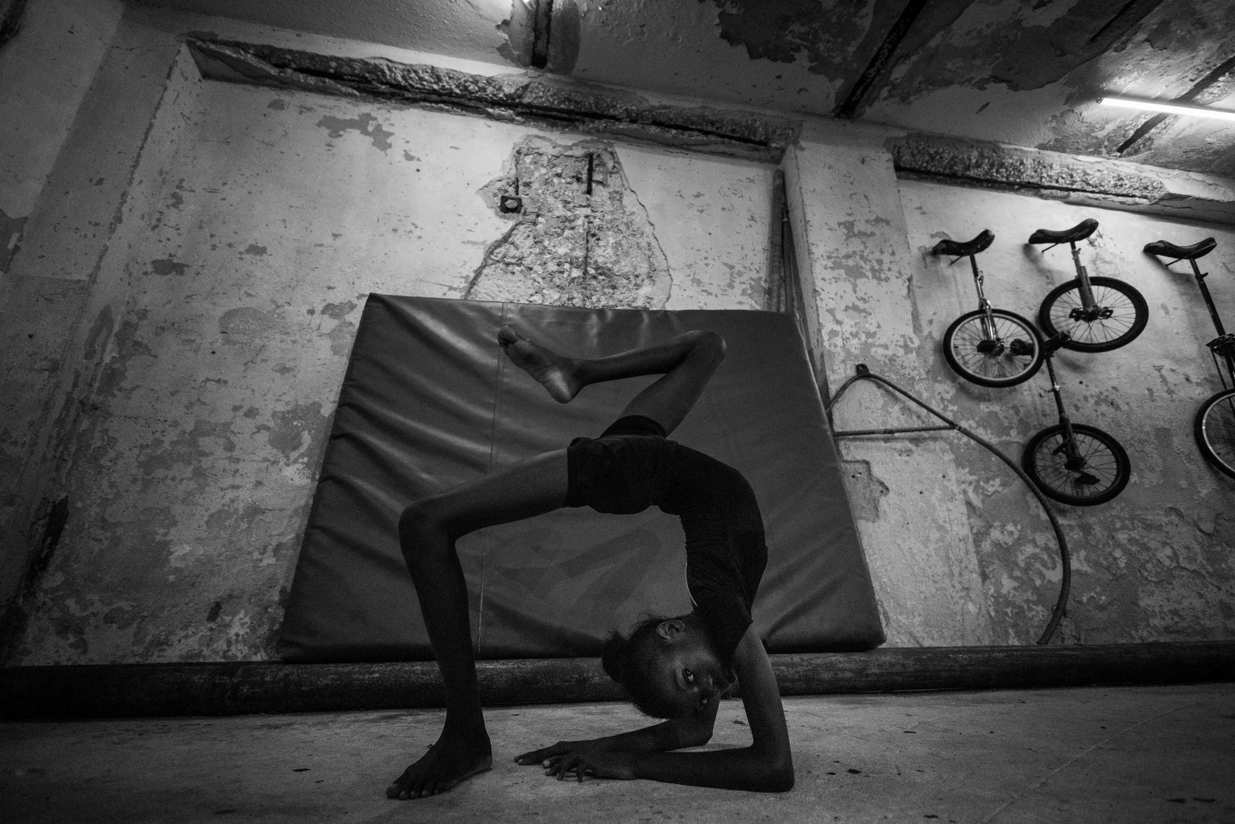 A young girl practicing a backbend yoga pose in a dimly lit, industrial-style room with peeling paint on the walls, three bikes hanging on the wall, and a wall-mounted hose.