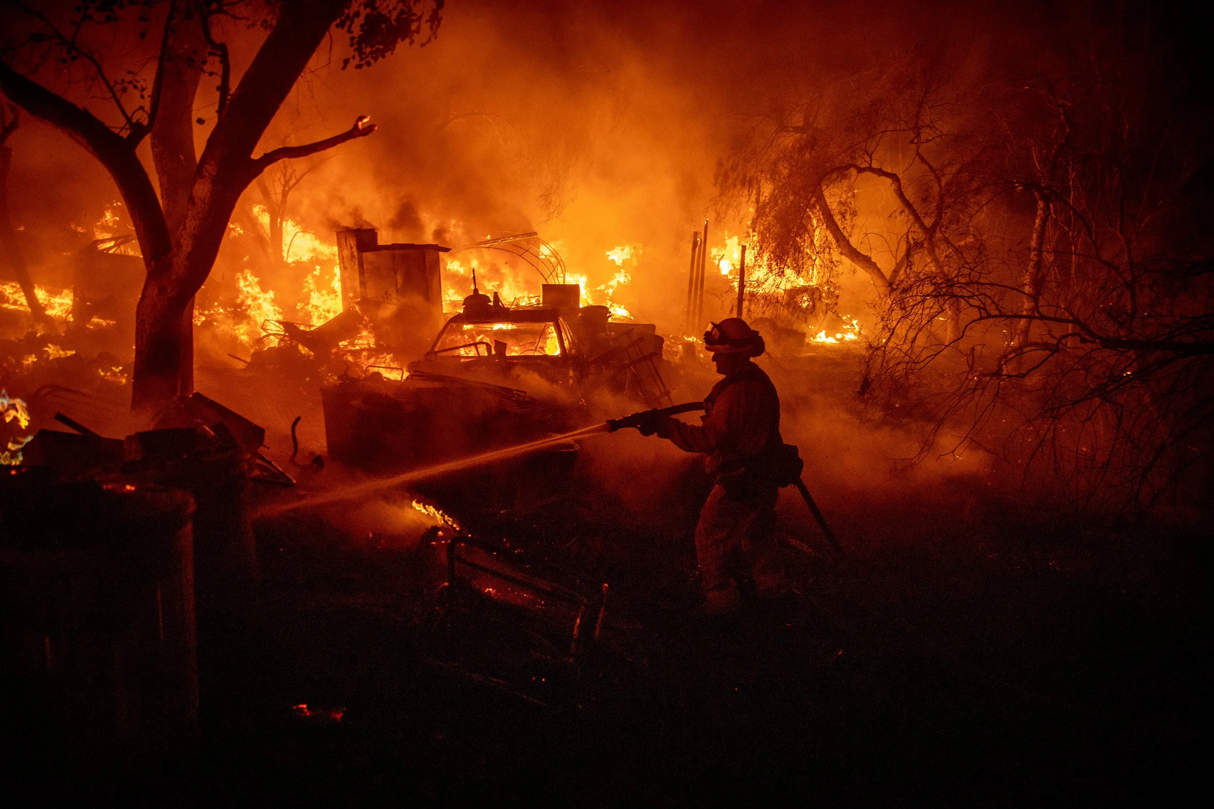 A firefighter in protective gear sprays water at a large house fire at night, with flames illuminating the background, charred trees, and smoke.