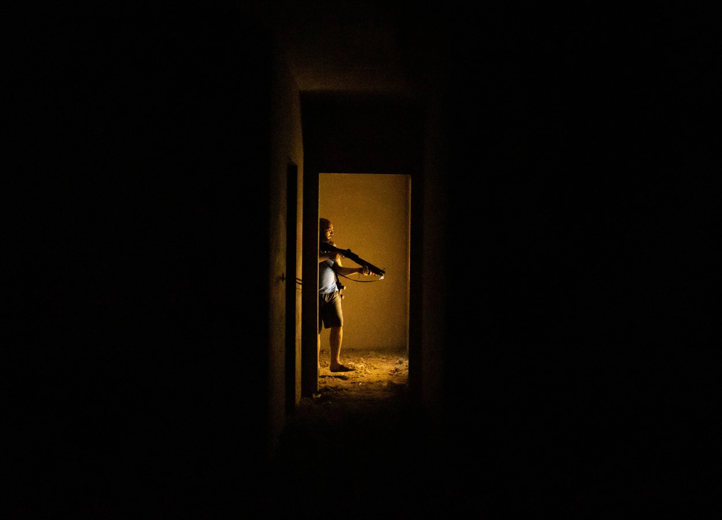 Person standing in a dimly lit doorway, holding a long weapon, with a yellowish glow illuminating the interior.