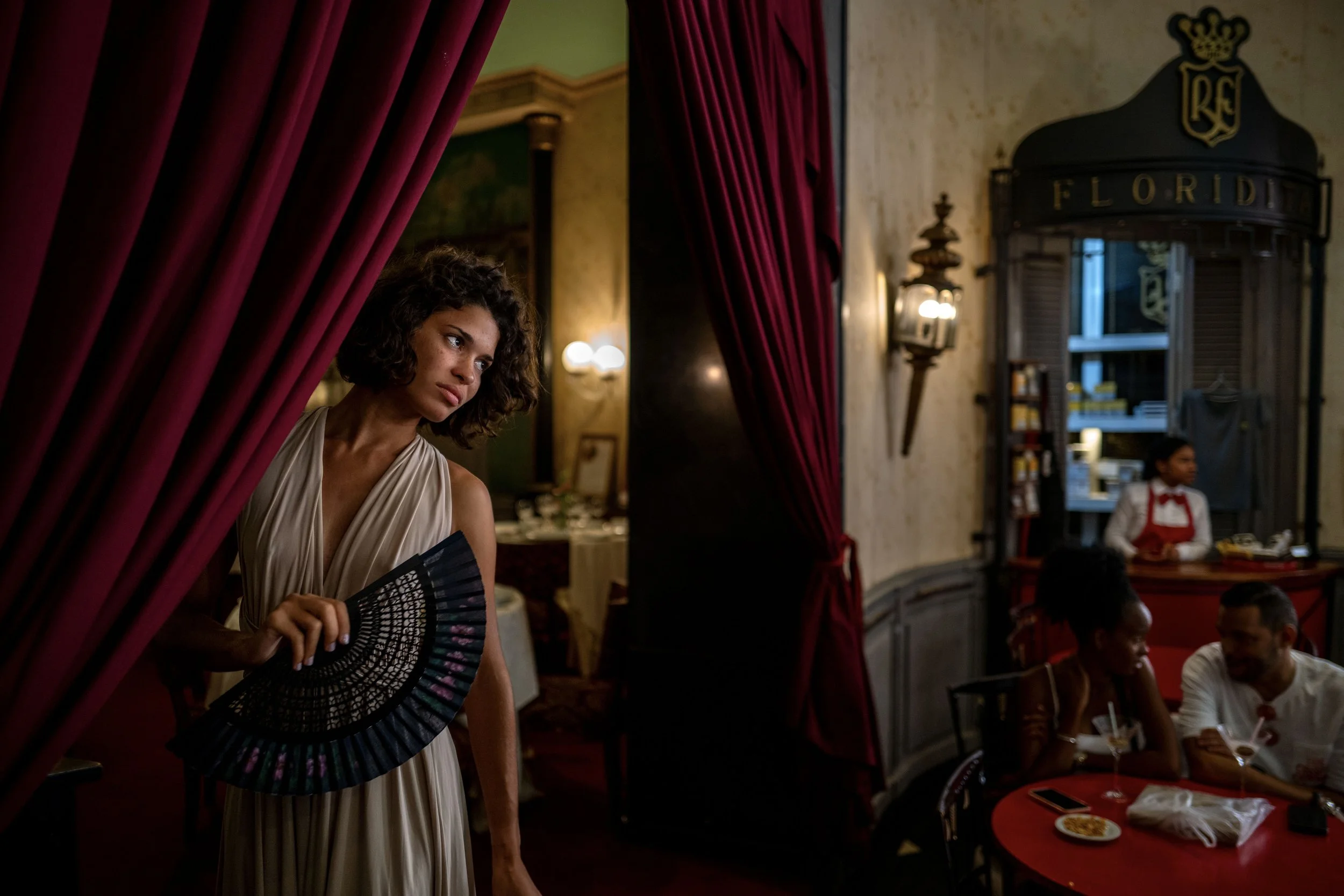 A woman in a beige dress holding a black fan peeks out from behind a red curtain inside a vintage-style restaurant, with decor and a server behind the counter in the background.