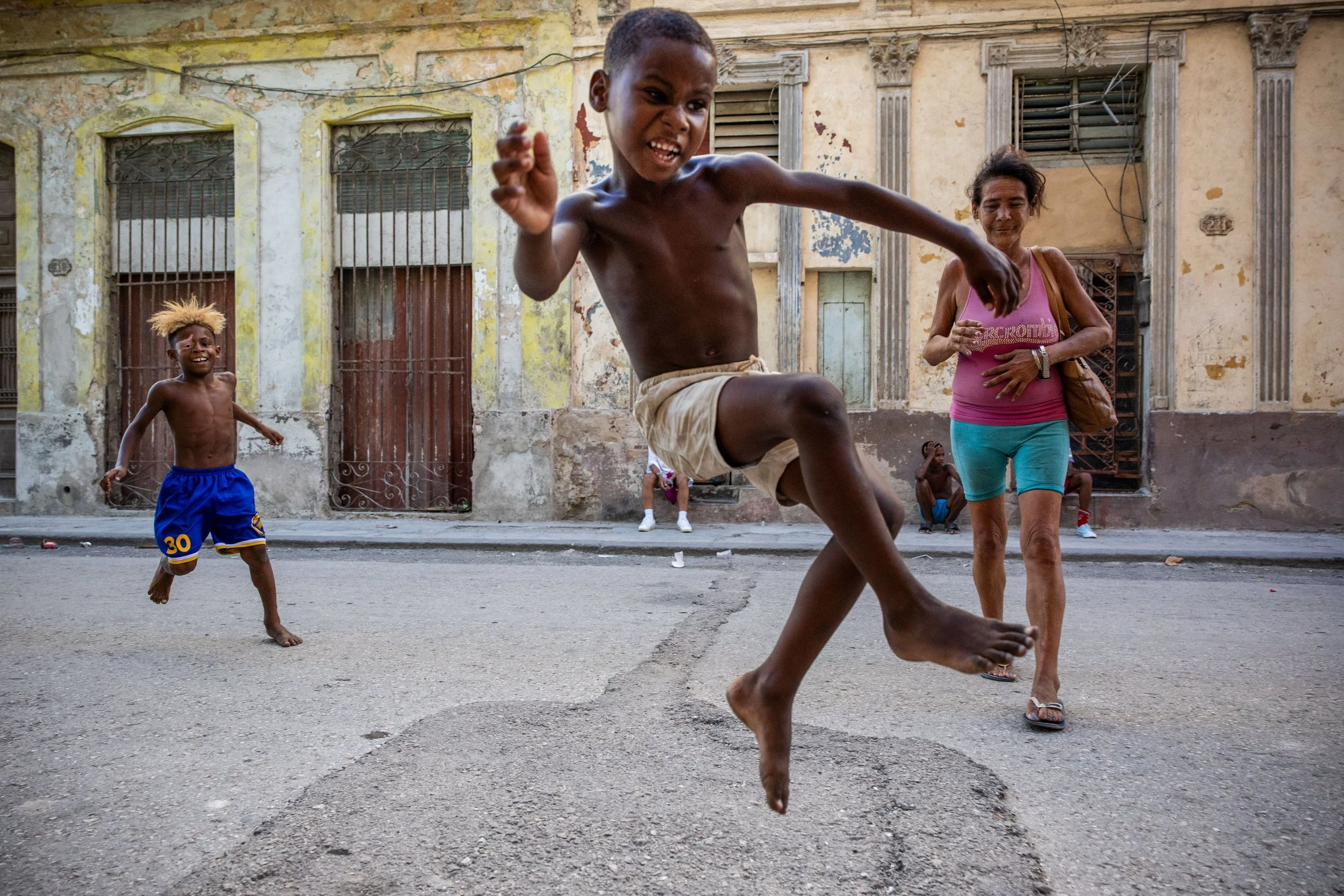 A boy jumps in the air with a joyful expression on a street, with a girl in blue shorts and a woman in pink and teal walking nearby. An older building with peeling paint and bars on windows forms the background.