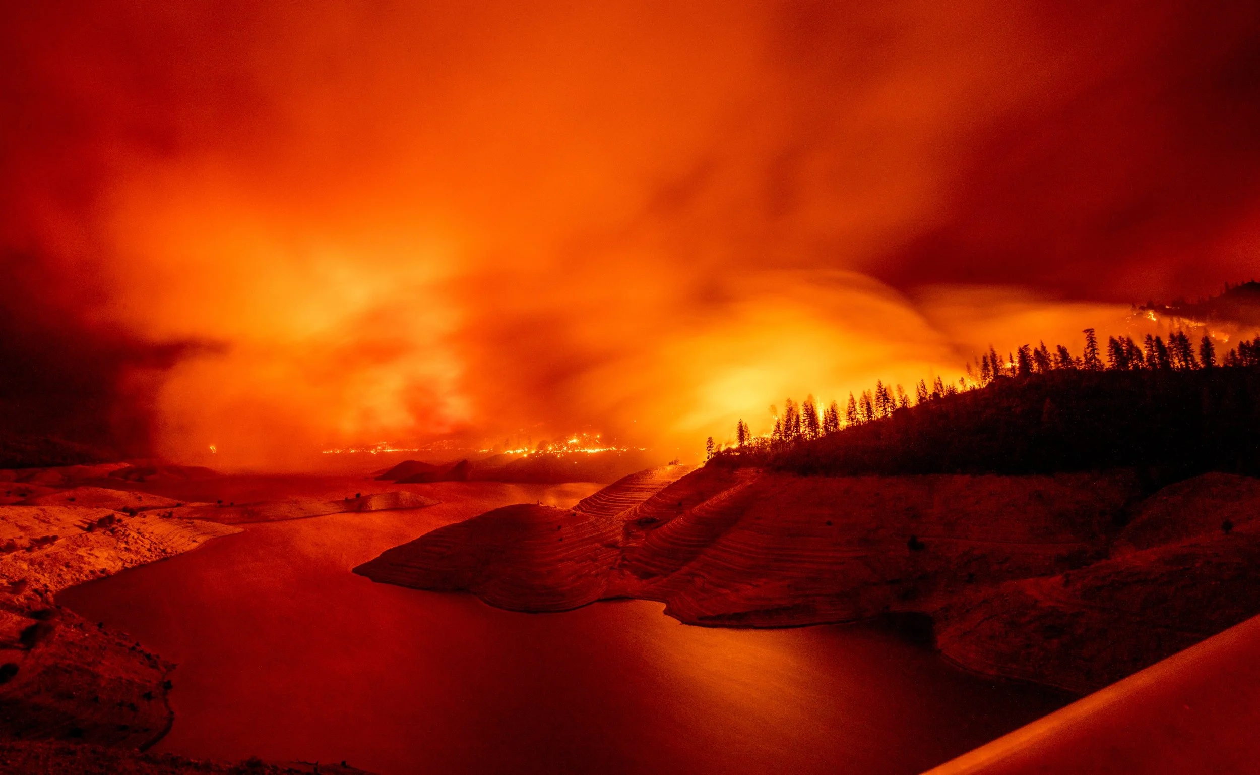 A landscape with a river flowing through terraced hills under a sky filled with dense, orange-illuminated smoke from a wildfire in the background.