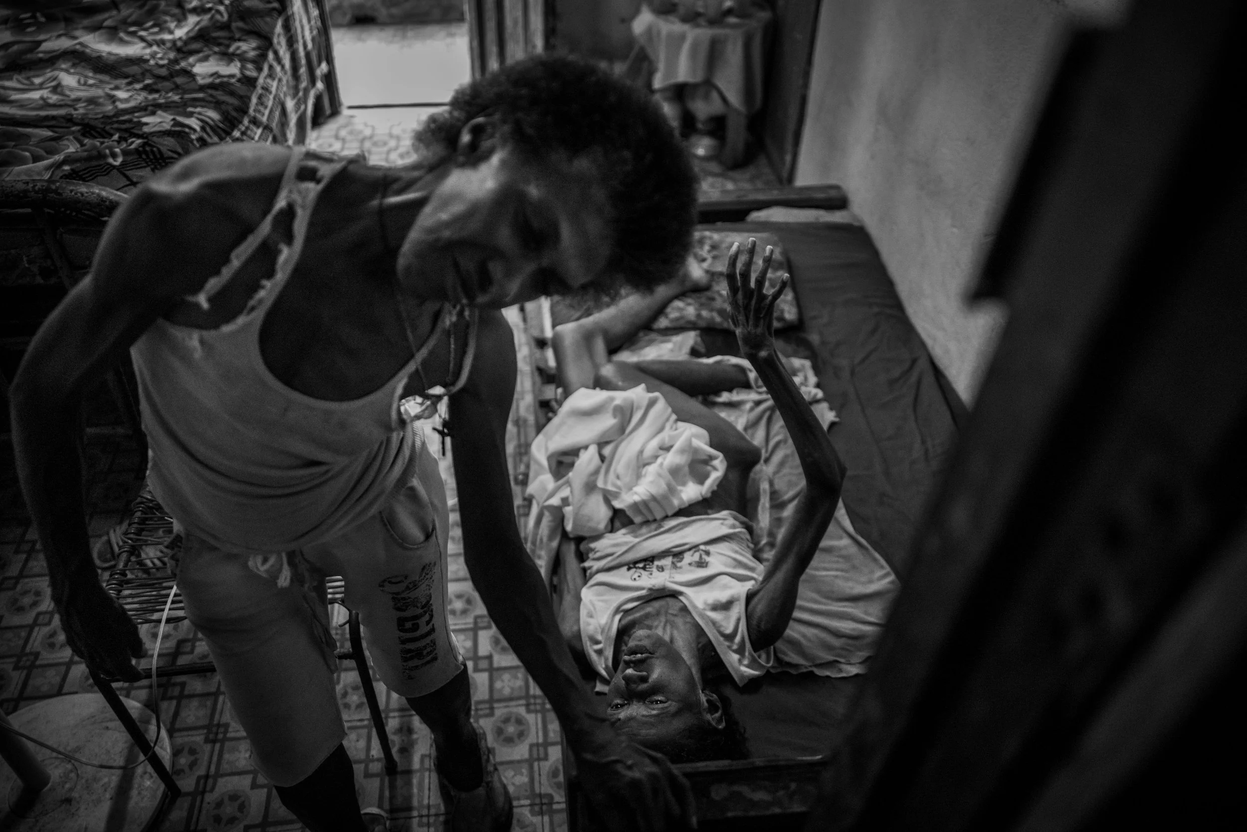 A distressed woman lying on a bed with a thin arm reaching toward her, as a man stands over her in a tense scene.