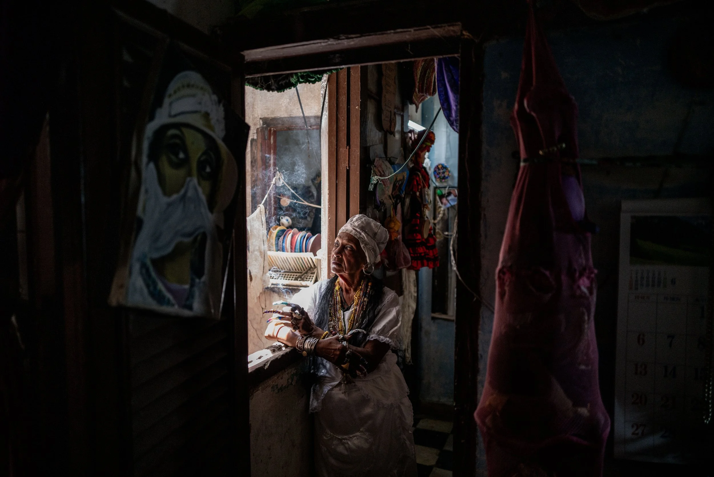 An elderly woman dressed in traditional clothing and jewelry, leaning on a window frame inside a dimly lit room, looking out.