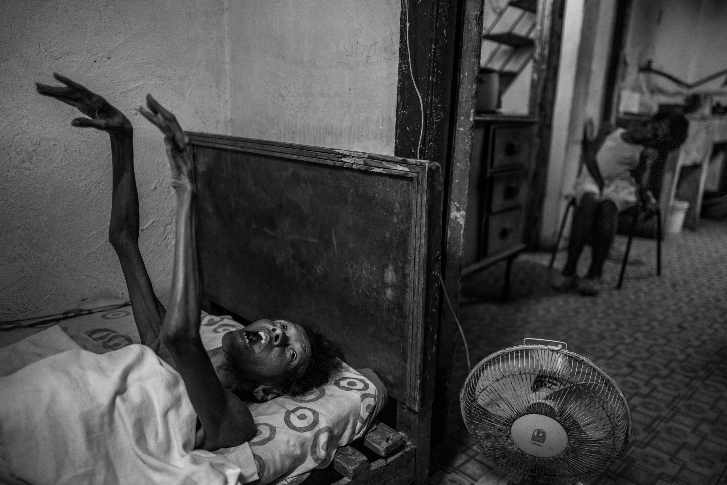 A black and white photo of a distressed woman lying in bed with her hands raised, in a modest room with a fan on the floor and another woman sitting on a chair in the background.