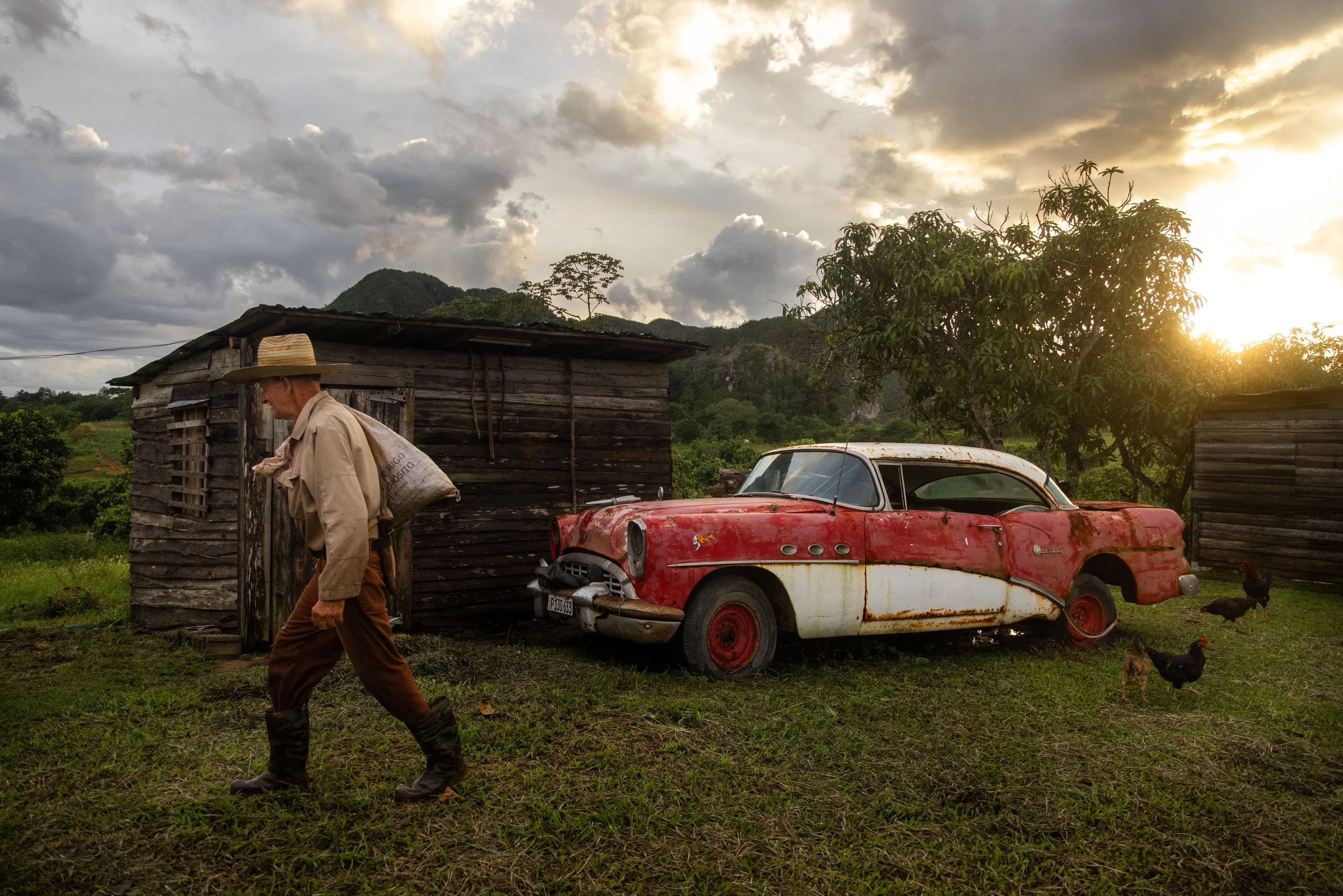 An elderly man wearing a straw hat, beige shirt, and brown pants walking past an old rusty red and white vintage car in a rural area during sunset, with wooden structures, chickens, trees, and mountains in the background.