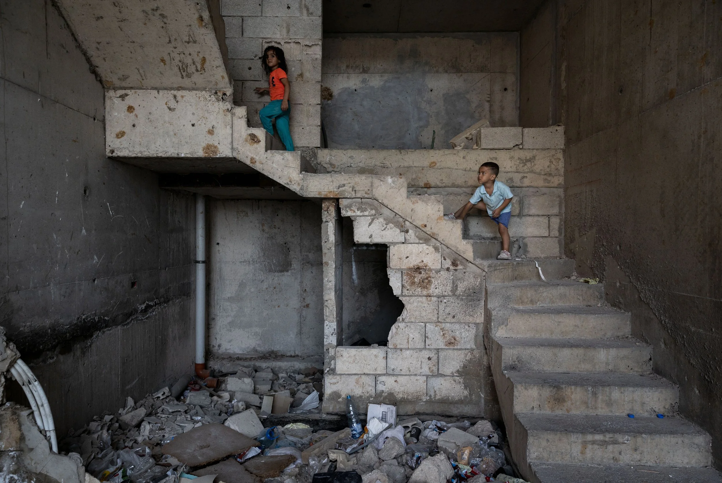 Two children playing on unfinished and partially damaged concrete staircase in a building under construction or and abandoned structure, with debris and trash on the floor.