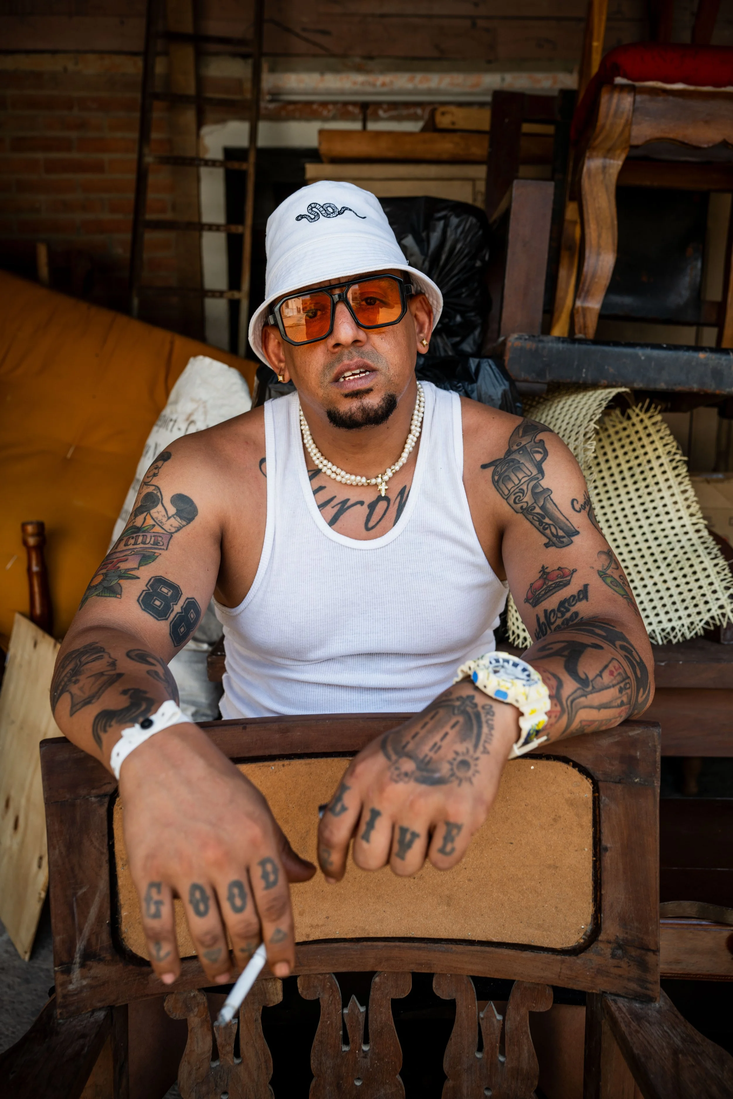 A tattooed man wearing a white tank top, sunglasses, a white bucket hat, and jewelry, sitting on a wooden chair in a rustic setting with wood and brick background.