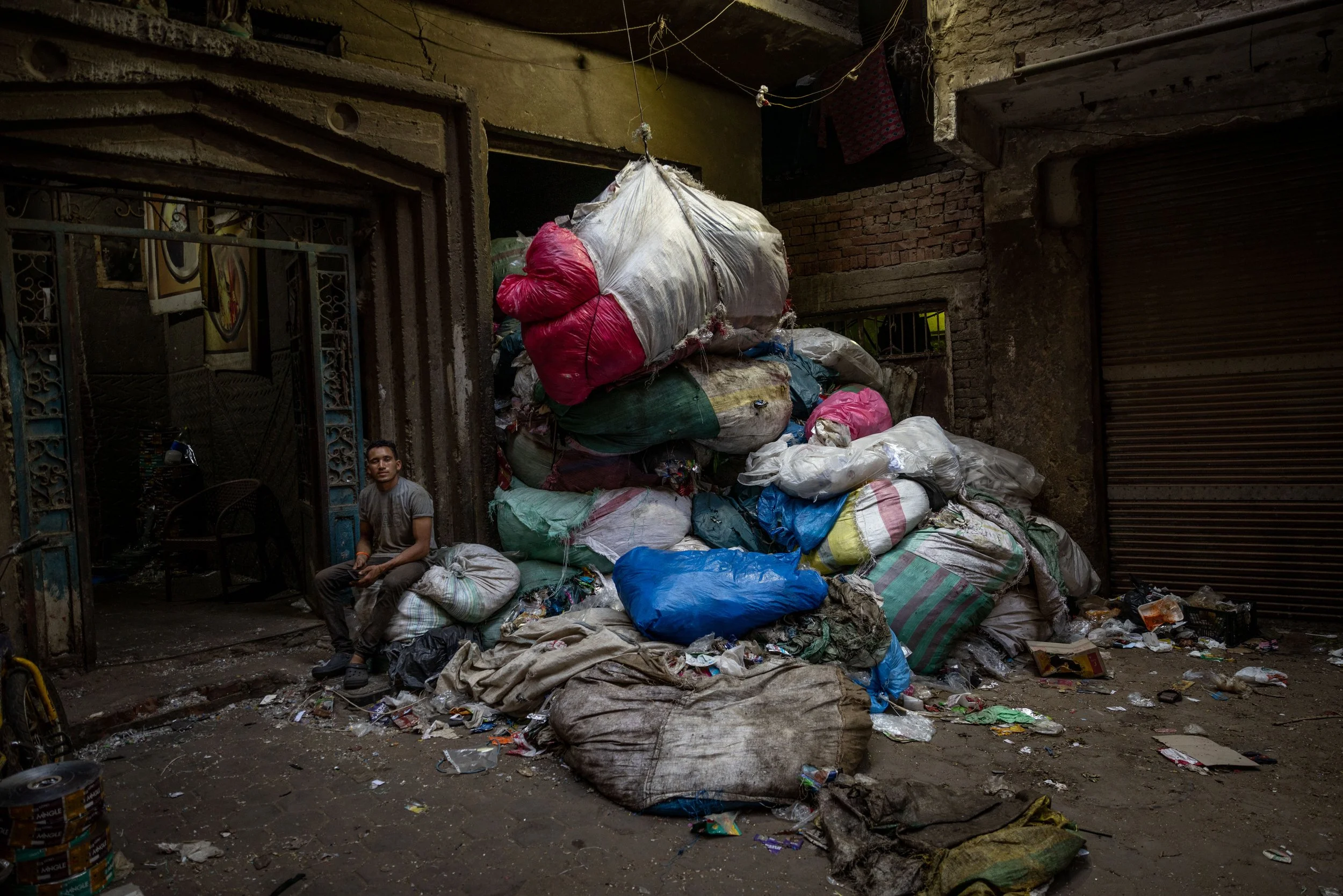 A young man sitting on a pile of garbage bags and trash outside a building with a large pile of dumped trash nearby.