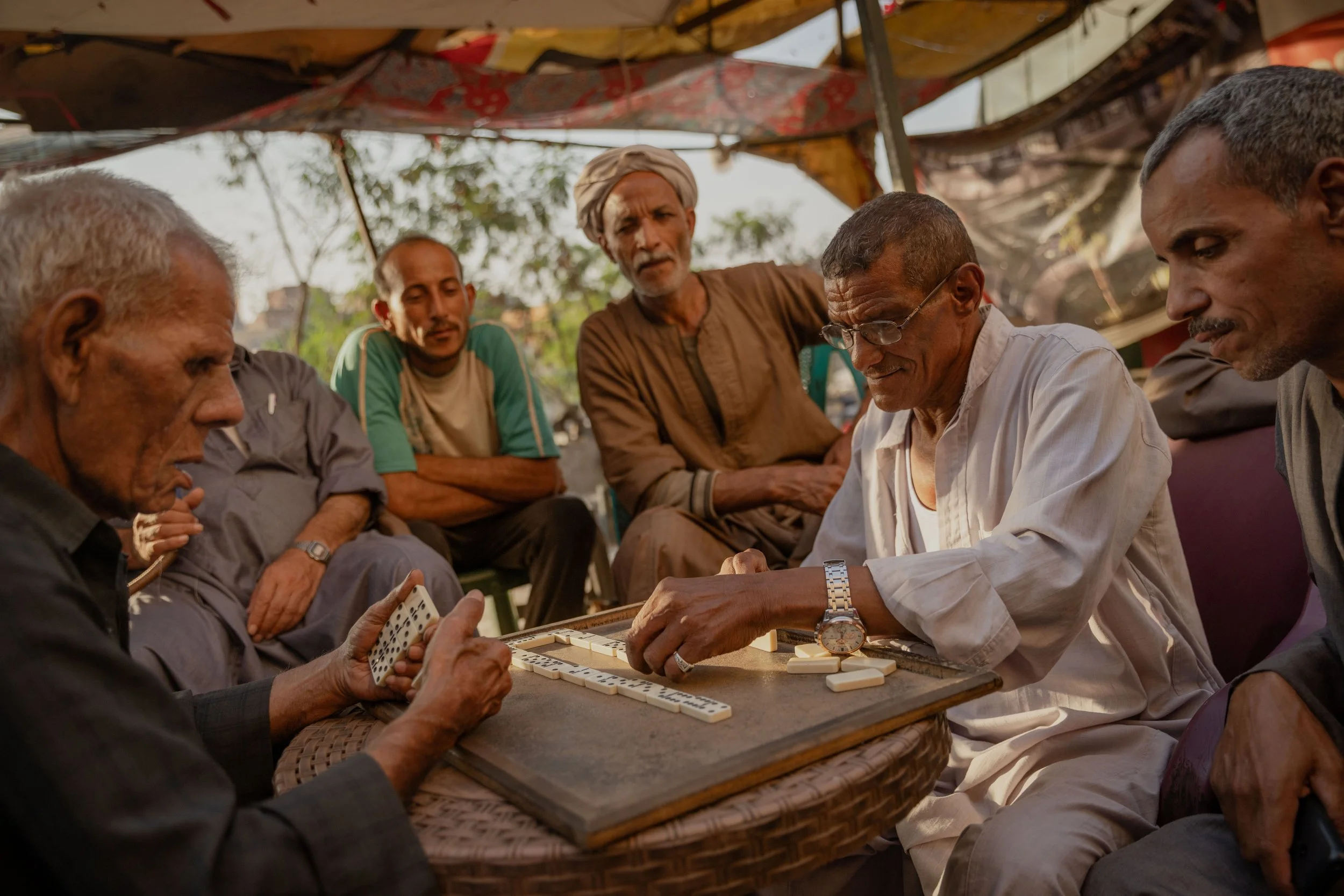 A group of six men playing dominoes outdoors under a canopy on a sunny day.