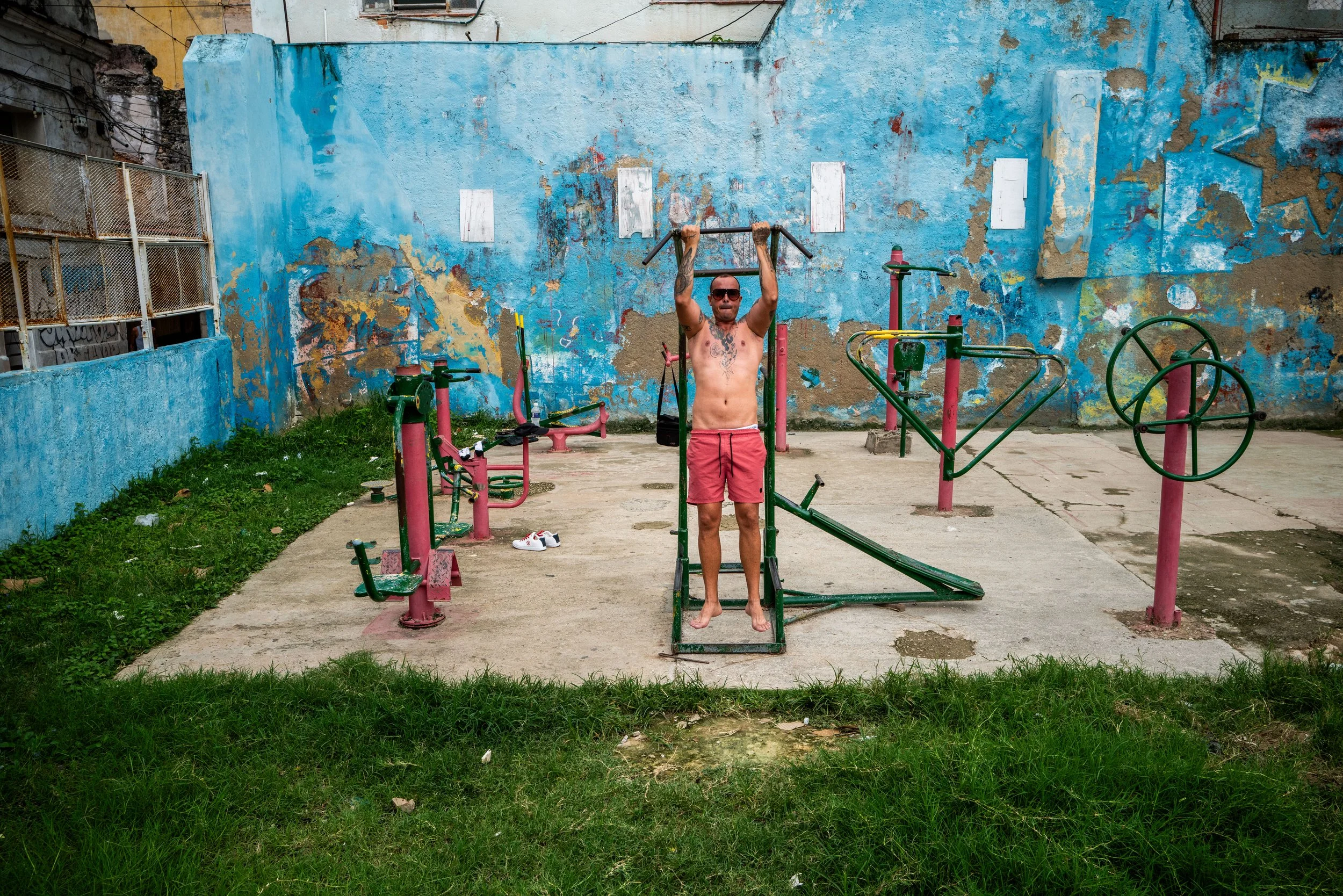 A man with sunglasses, tattoos, and wearing red shorts, is shirtless and exercising on outdoor gym equipment in a rundown park with a blue peeling wall in the background. He is holding onto a pull-up bar.