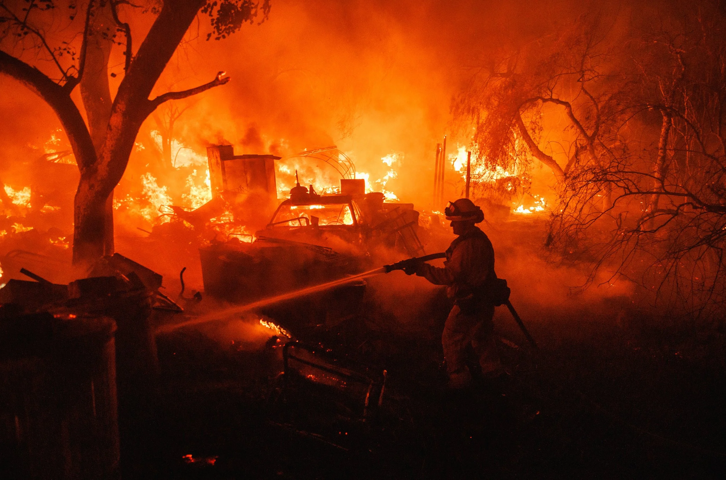 A firefighter in protective gear spraying water on a large house fire at night, with bright flames, smoke, and burning trees surrounding the scene.