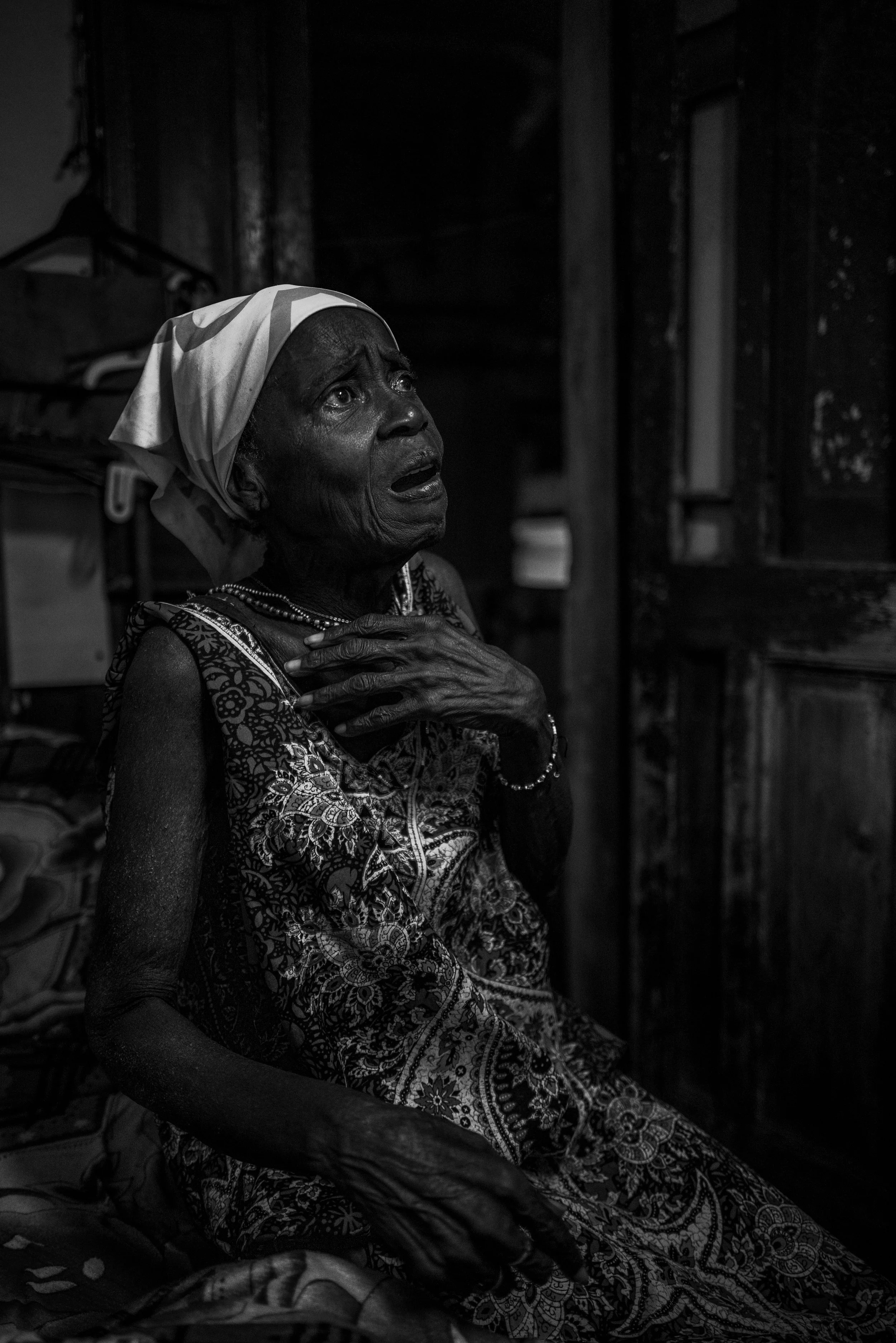 An elderly woman with a distressed expression, sitting indoors, wearing a patterned dress and a headscarf, with her hand on her chest.