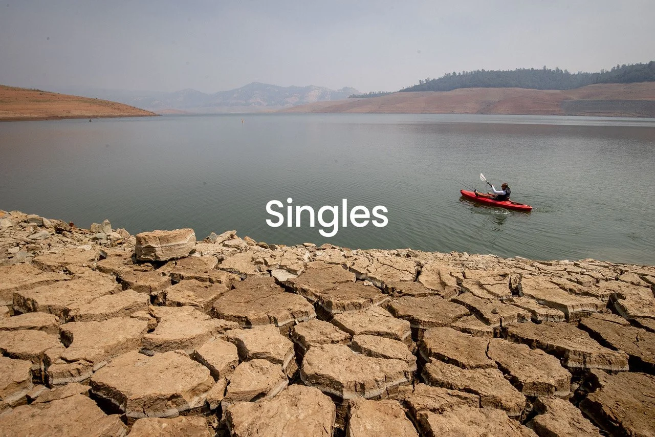 Person kayaking on a large body of water with dry, cracked earth in the foreground and hills in the background, overlaid with the word 'Singles'.