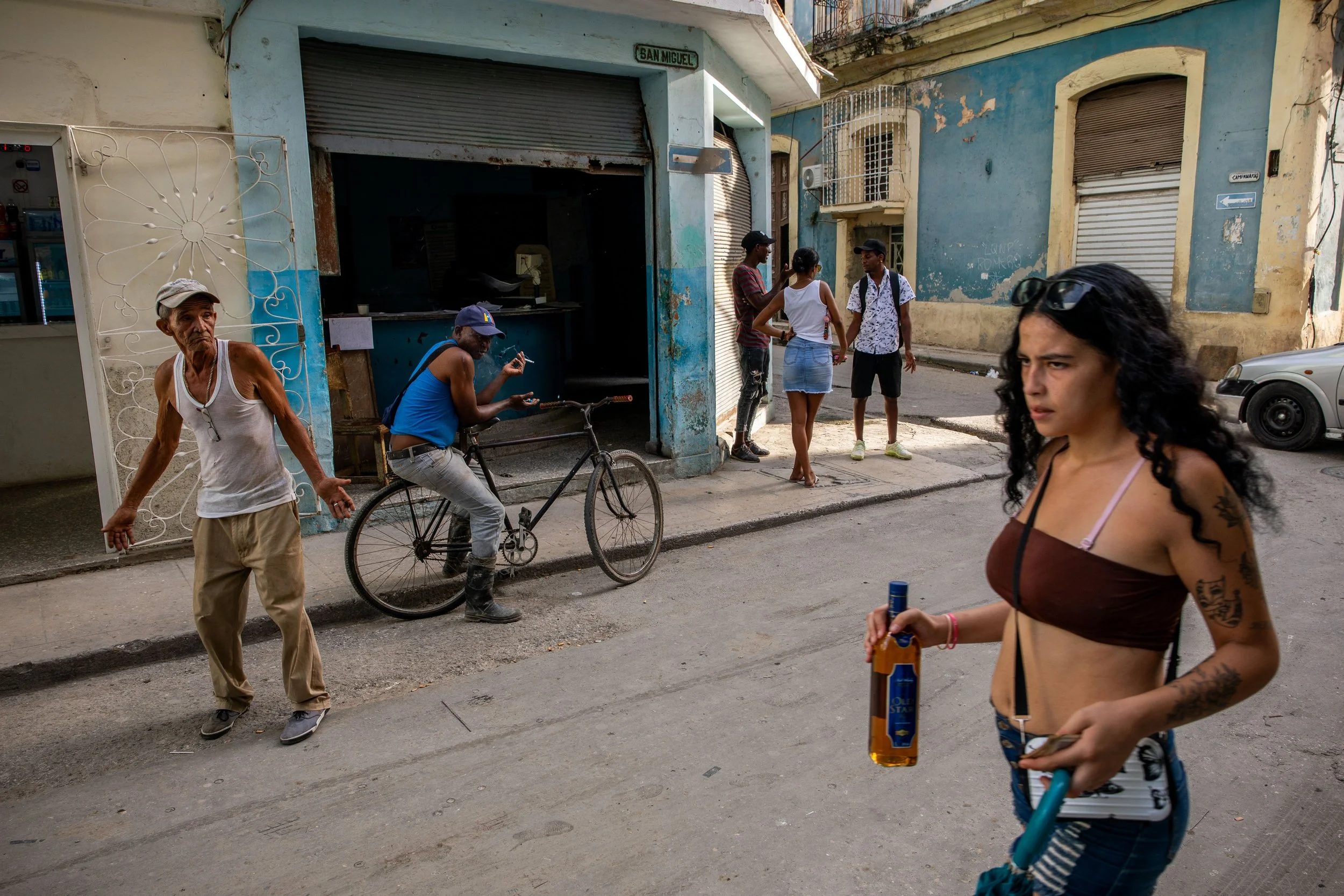 Street scene with five people: an older man in a white tank top and beige pants standing on the sidewalk, a young man in a blue tank top and gray pants sitting on a bicycle, a group of three young people talking near a blue building, and a young woma