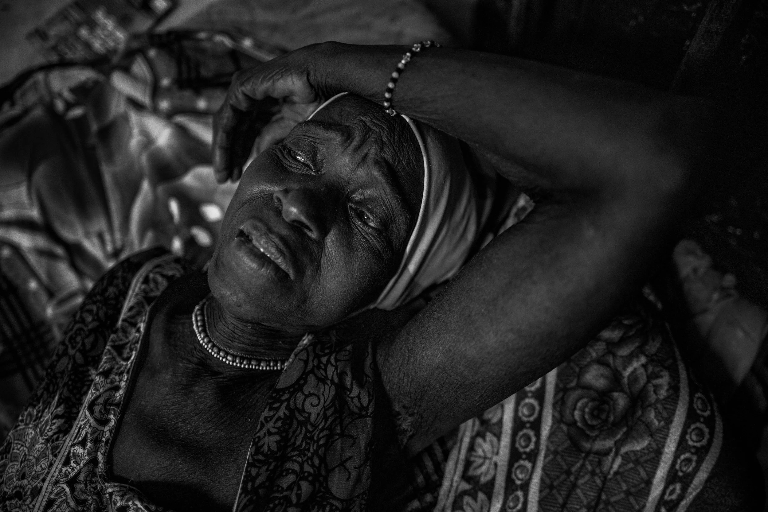 A black and white photo of an elderly woman lying down, resting her head on her arm, with her eyes closed and a peaceful expression on her face, wearing traditional jewelry and patterned clothing.