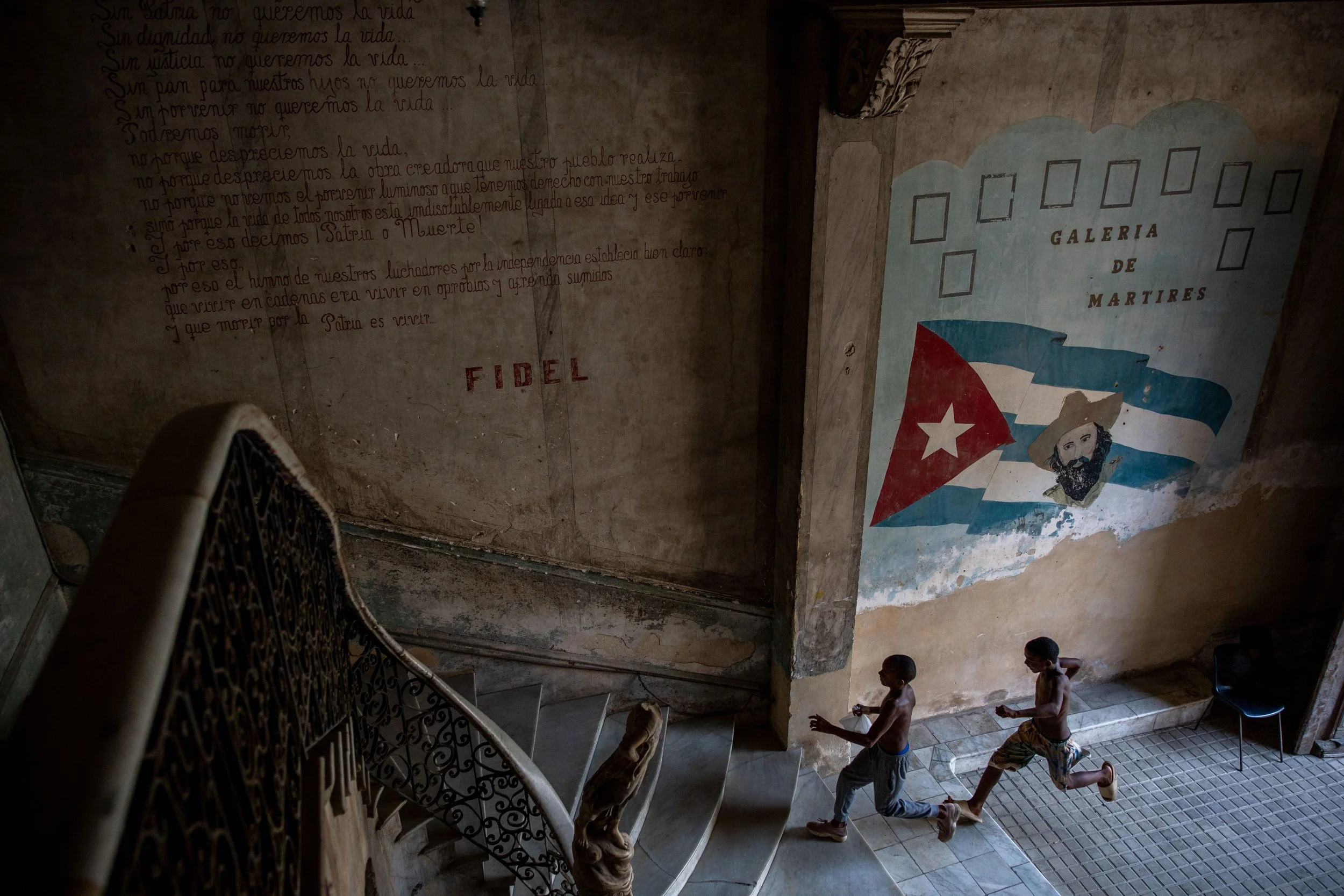 Two young boys run down a staircase inside a building with worn walls. On the wall to the right, there is a mural of the Cuban flag with a portrait of a man in a hat. To the left, handwritten Spanish text is visible, along with the word 'FIDEL' writt