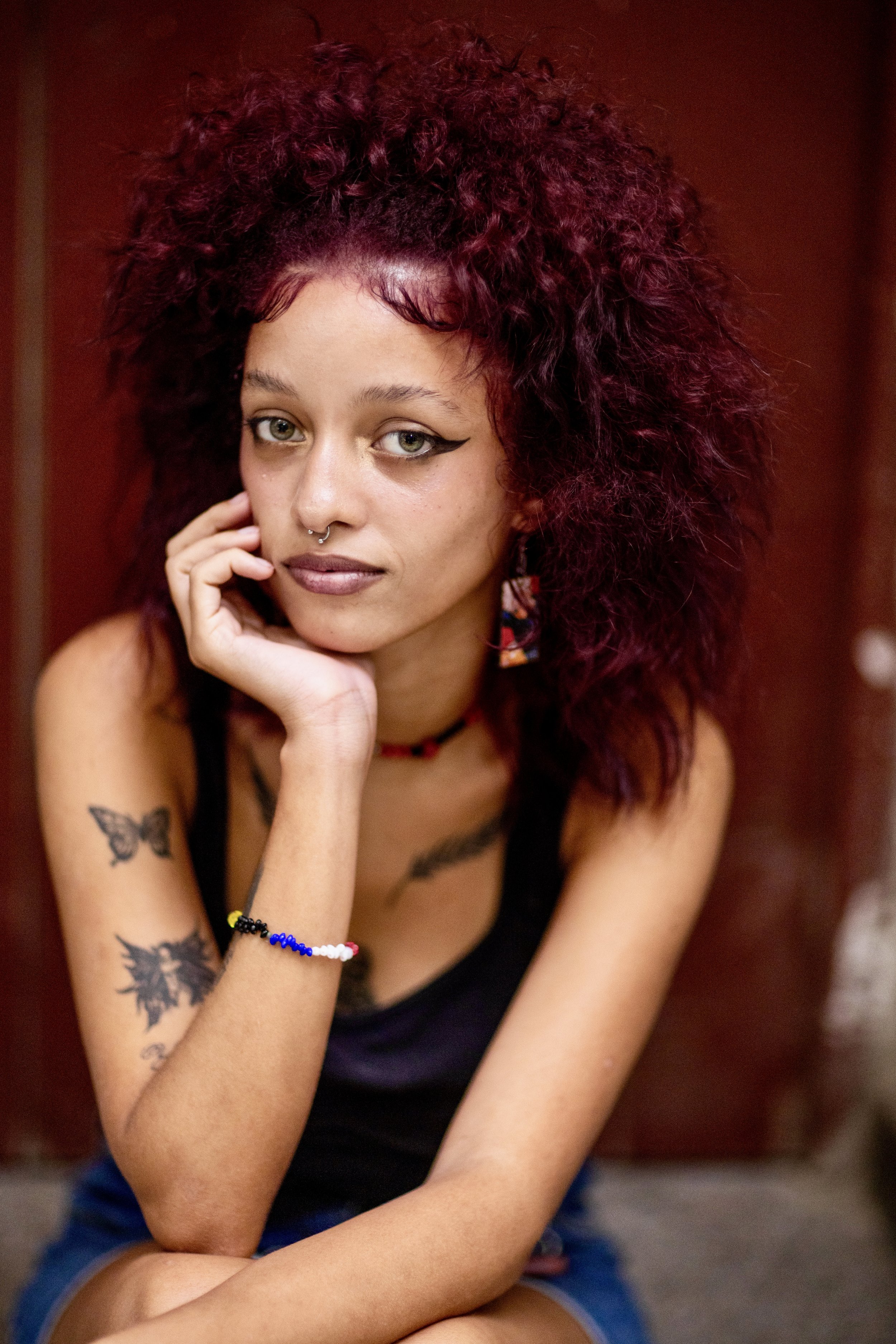 A young woman with curly, reddish hair, wearing a black tank top, colorful earrings, and a bracelet, sitting with her chin resting on her hand and looking at the camera. She has tattoos on her arm and a septum piercing.
