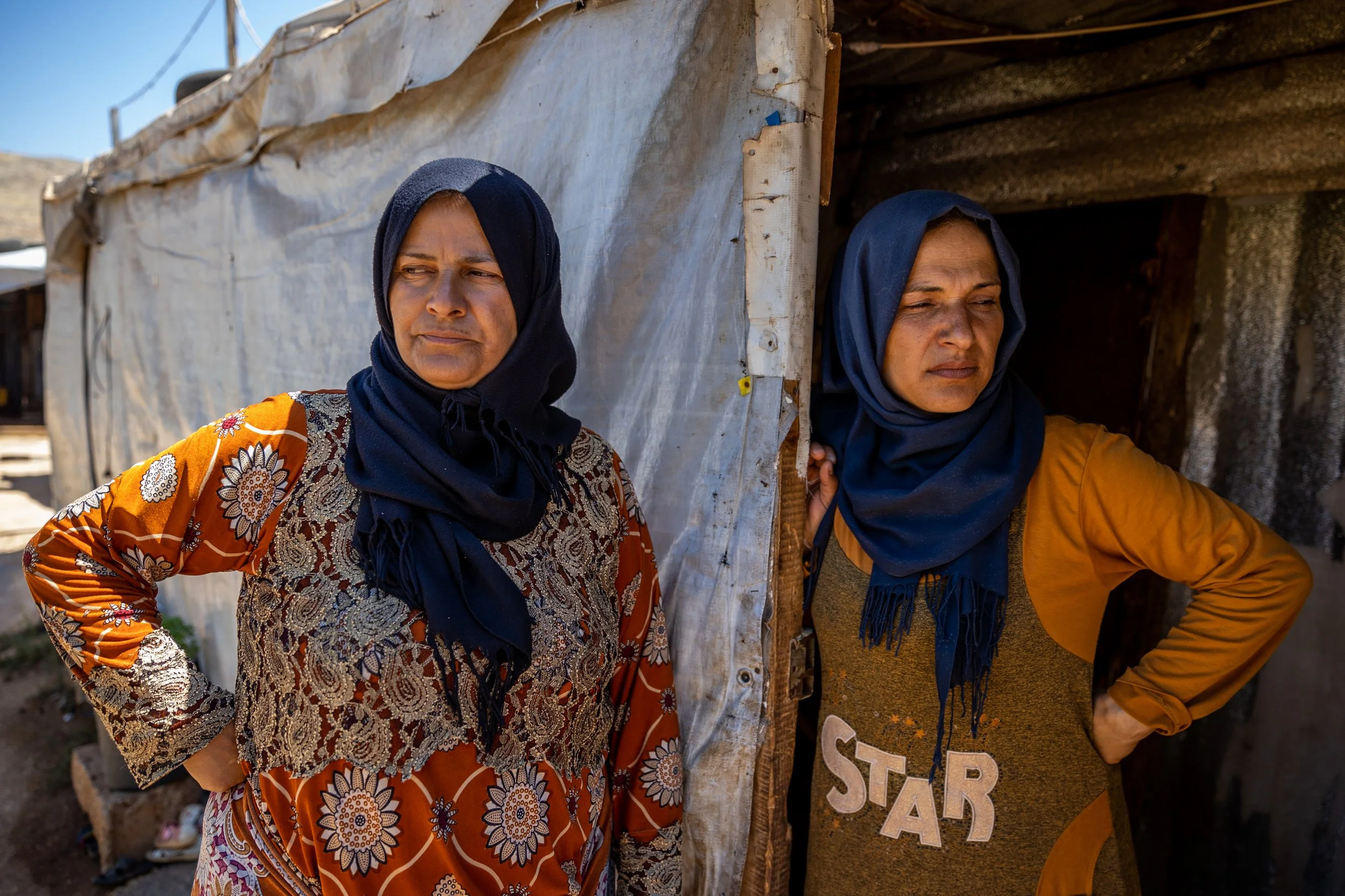 Two women wearing dark hijabs stand outside a makeshift shelter made of fabric and wood, with serious expressions.