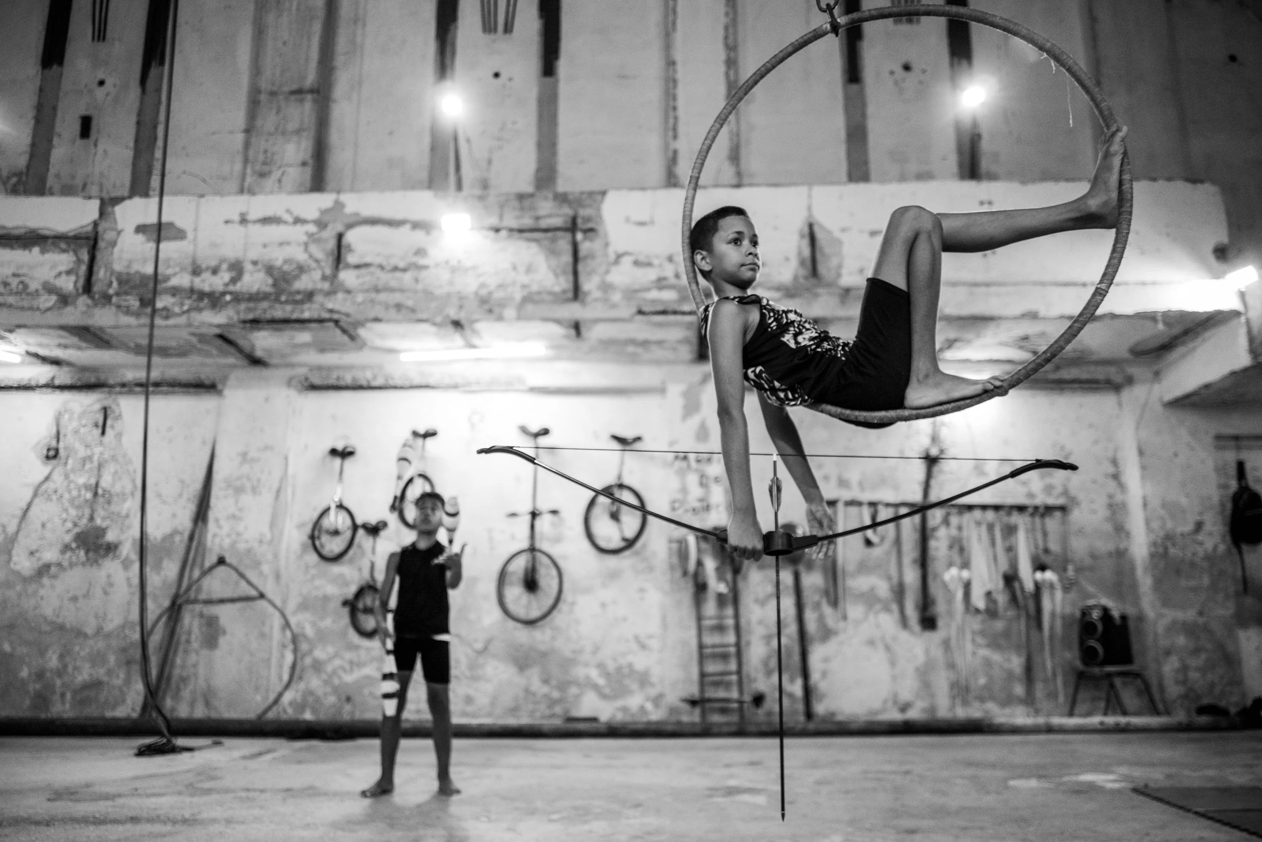A young girl performing on an aerial hoop inside a circus or gymnasium, with a boy standing in the background. The space has a rustic, worn look with hanging bicycles and equipment on the walls.