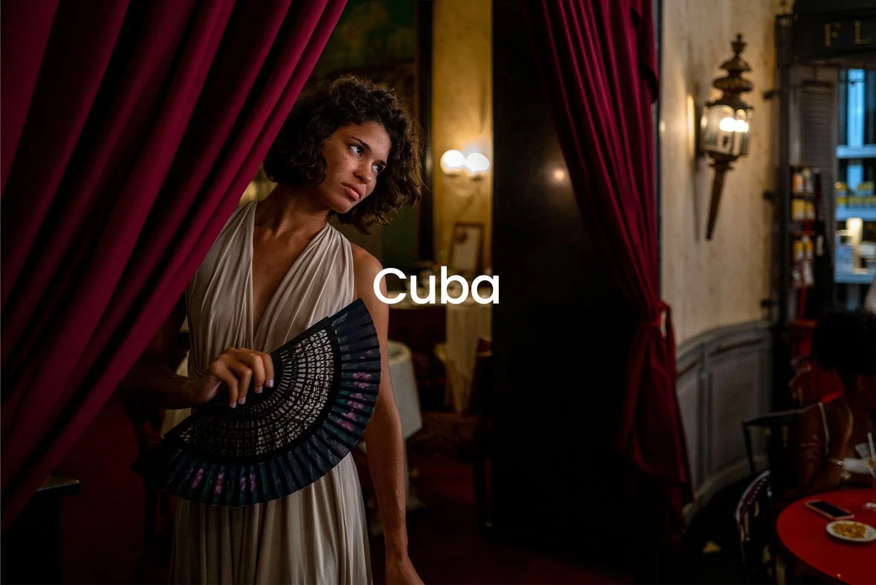 A woman with curly hair and a beige dress holding a black hand fan, standing in a dimly lit restaurant or café in Cuba, with red curtains on the sides and warm lighting.