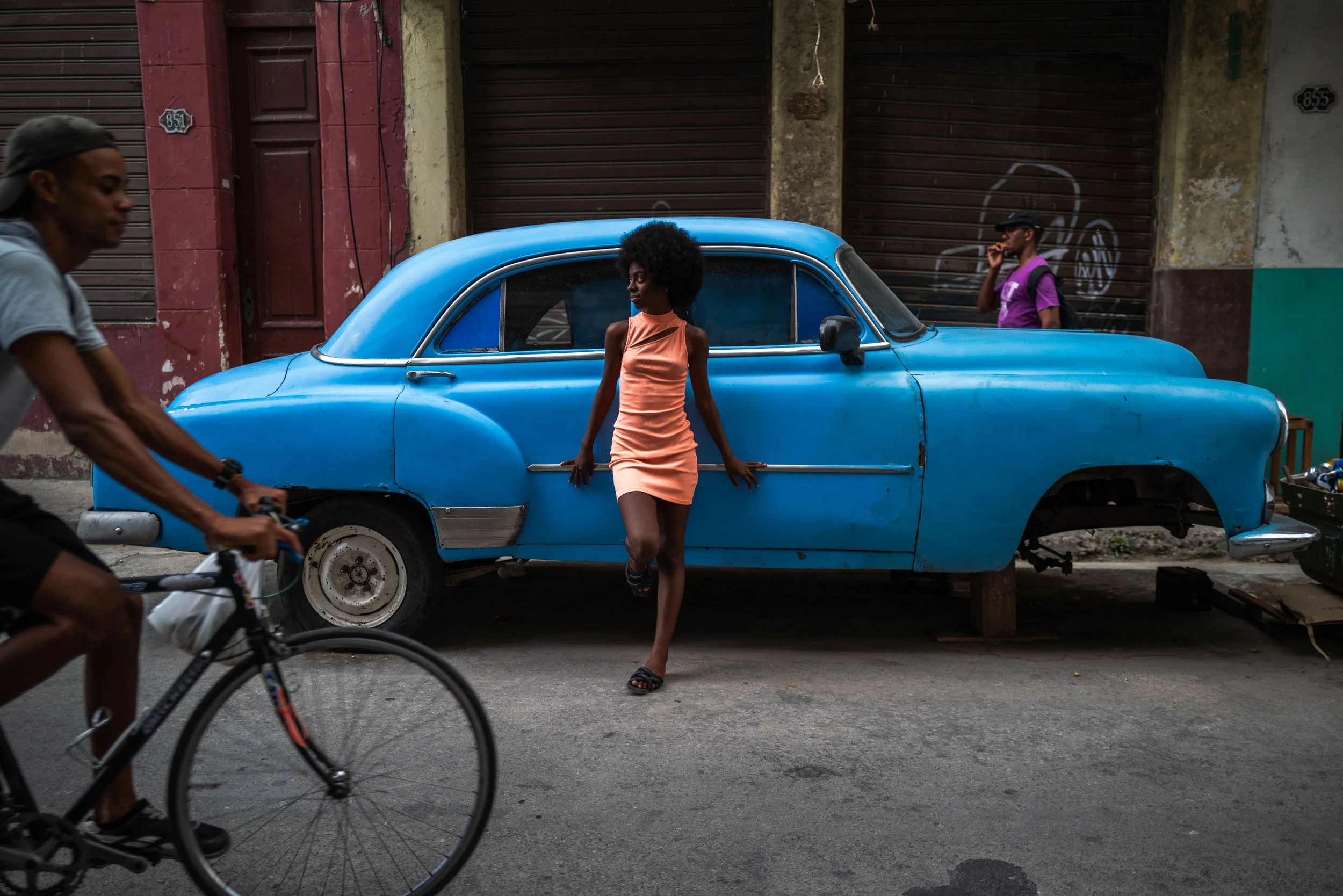 A woman in a peach dress leaning against a bright blue vintage car on a city street, with two men walking and talking in the background, and a cyclist passing by.