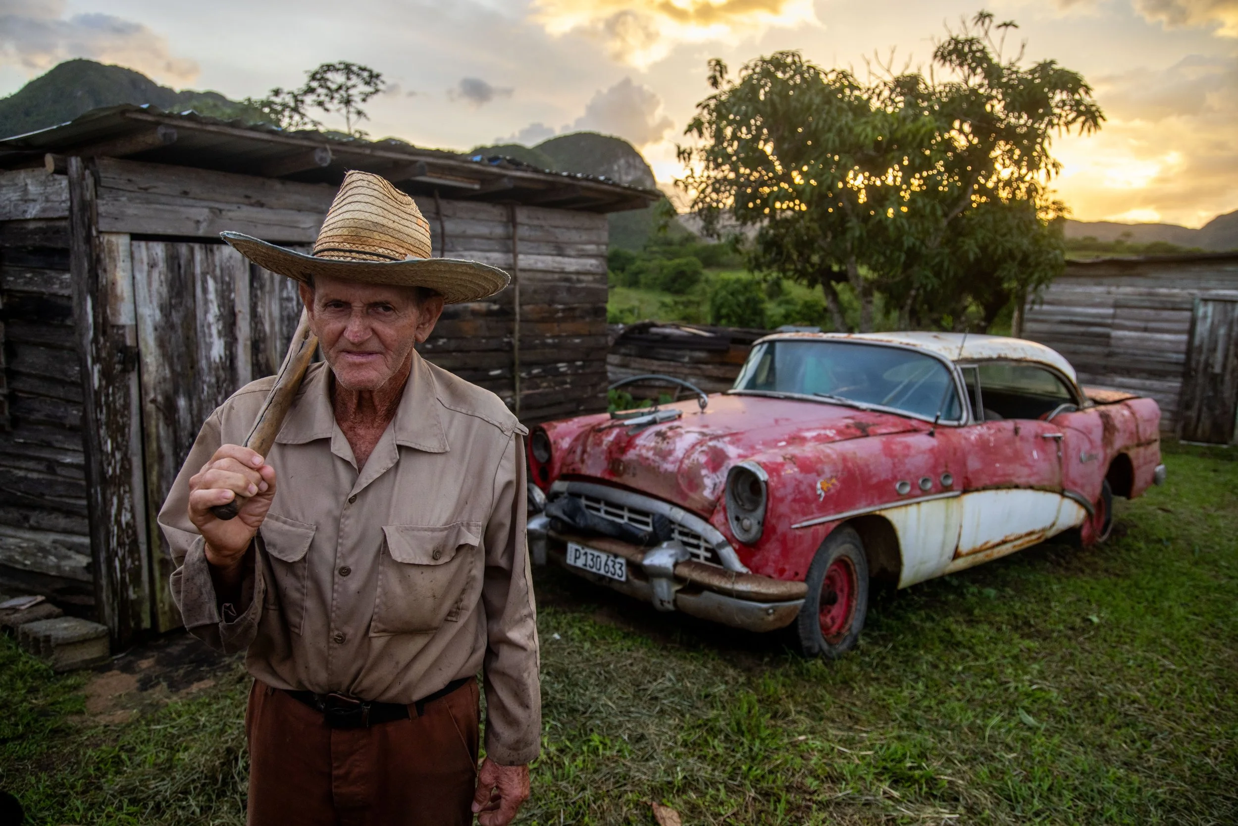 An elderly man wearing a straw hat and a beige shirt holds a walking stick, standing in front of an old, rusty red and white vintage car and weathered wooden shed, with trees and mountains and a sunset sky in the background.
