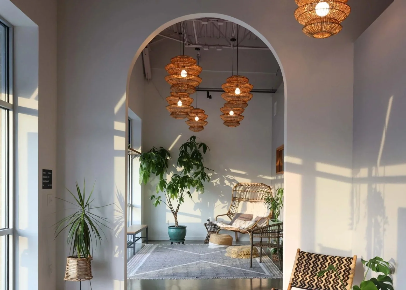Interior of a modern space with hanging pendant lights, large windows, indoor plants, wicker furniture, and a chalkboard sign with colorful writing.