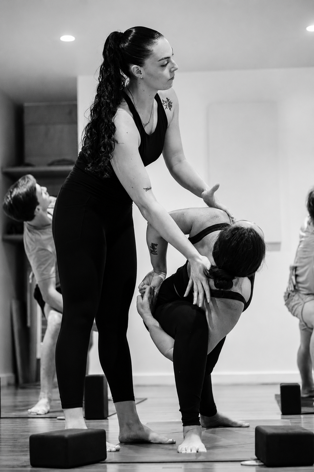 Two women practicing yoga, with one woman assisting the other in a supported forward bend, on mats in a studio.