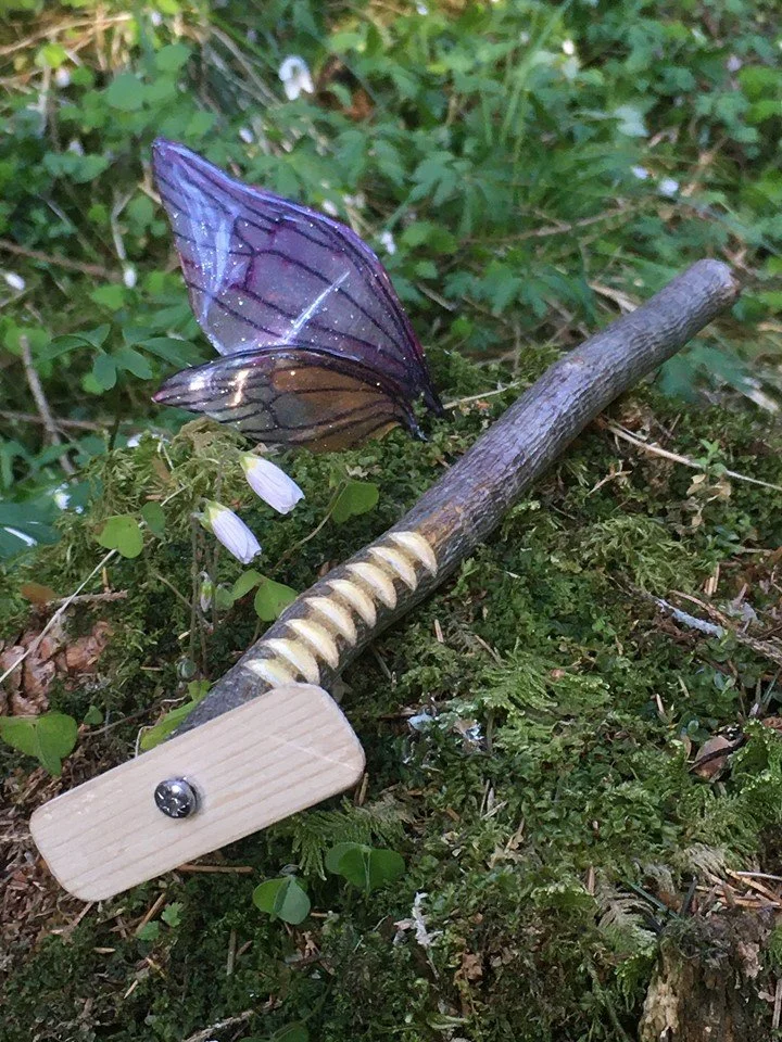 A toadstool mushroom with a white cap growing among green moss and small plants. A handmade wooden stick with a screw and a fitted handle is placed nearby on the ground.