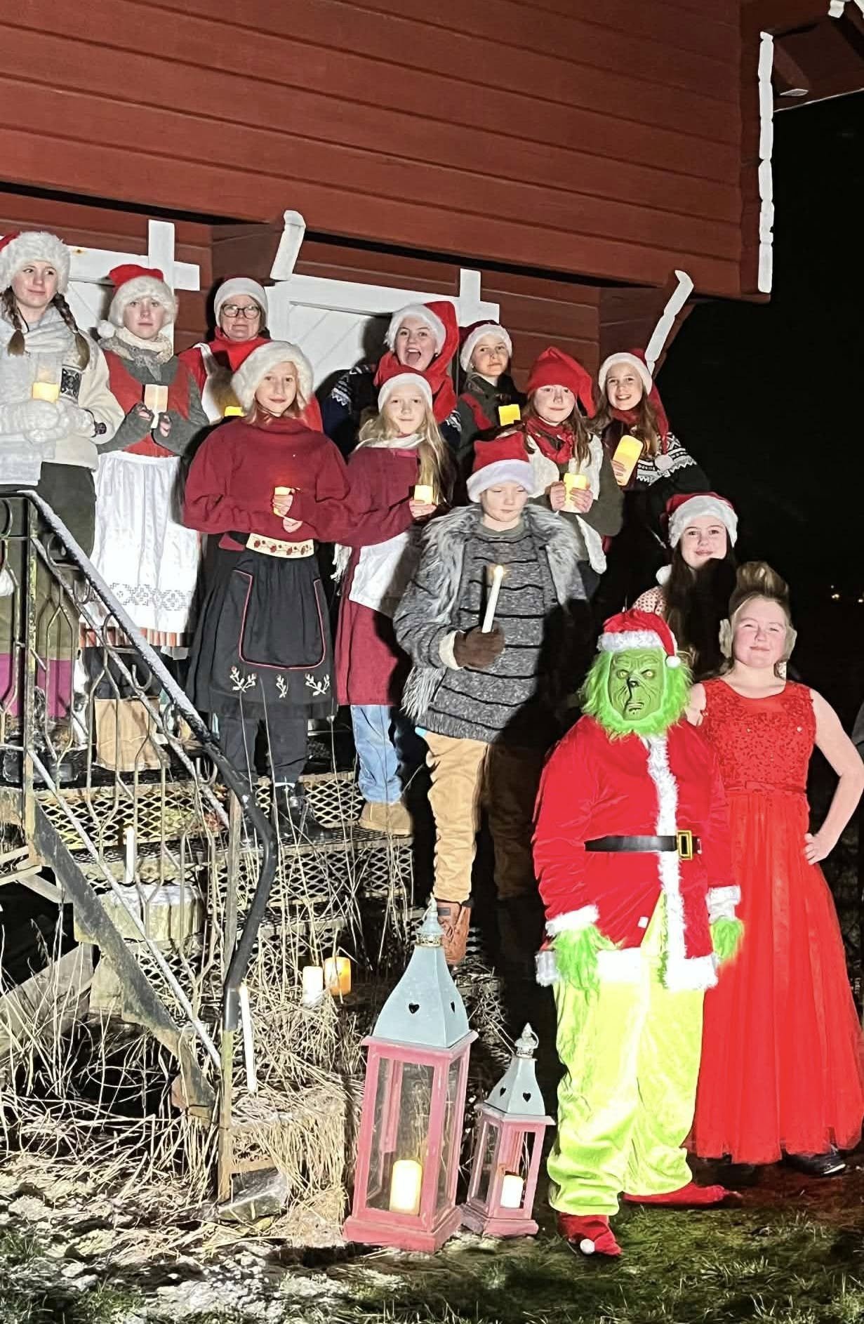 Group of children and two adults dressed in Christmas costumes, including Santa Claus and the Grinch, standing on a porch with Christmas candles and decorations at night.