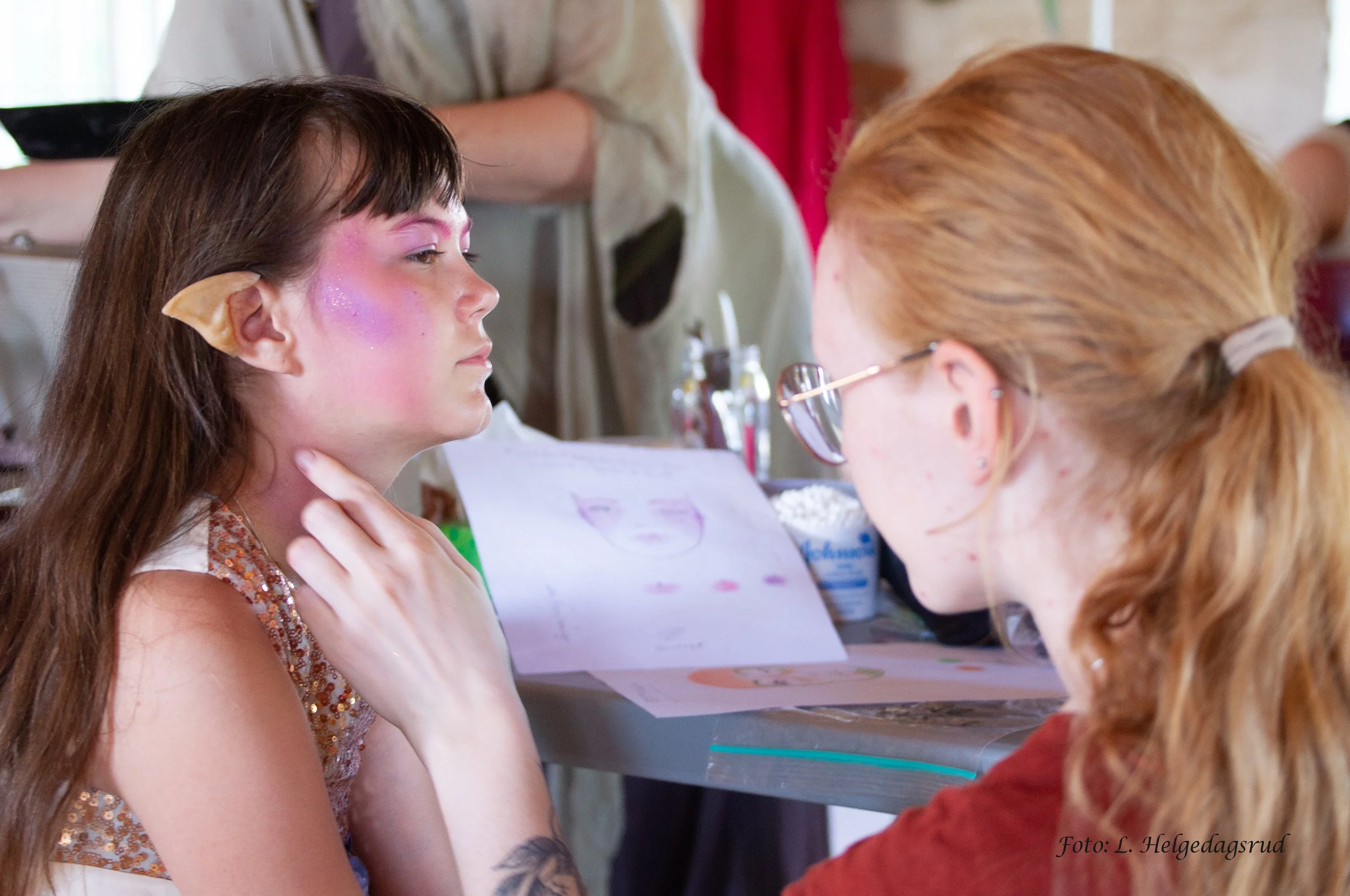 An artist is applying makeup or face paint on a girl with elf-like features, including pointy ears, at a face painting event. The girl has colorful makeup with pink and purple hues, and the artist has red hair tied back, wearing glasses. There are art supplies and drawings on the table.