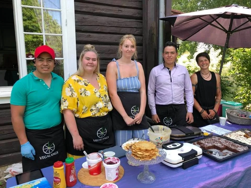 Five people standing behind a table with food at an outdoor event, with a wooden building and trees in the background.
