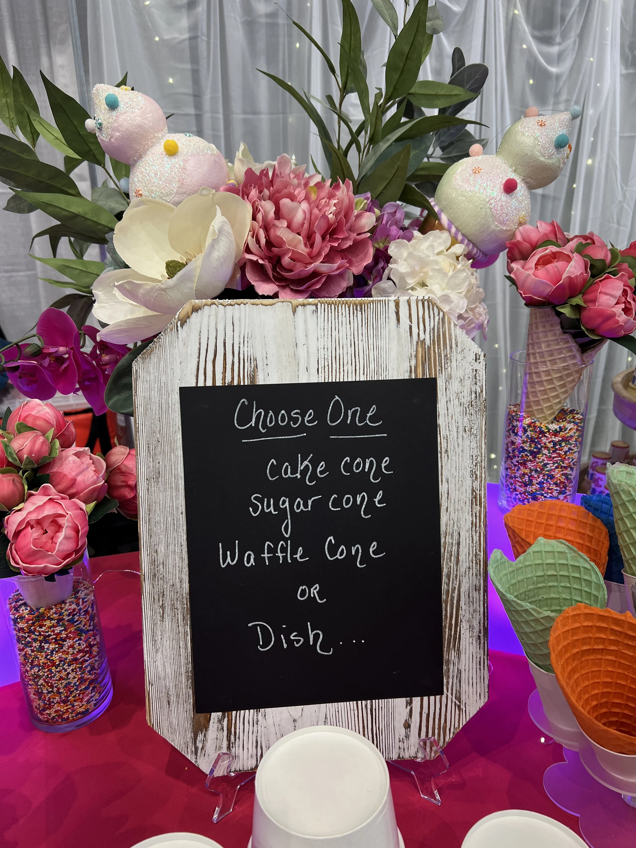 Decorative floral display with cotton candy shaped like clouds, surrounded by pink, white, and purple flowers, and colorful sprinkles in glass jars and ice cream cones.