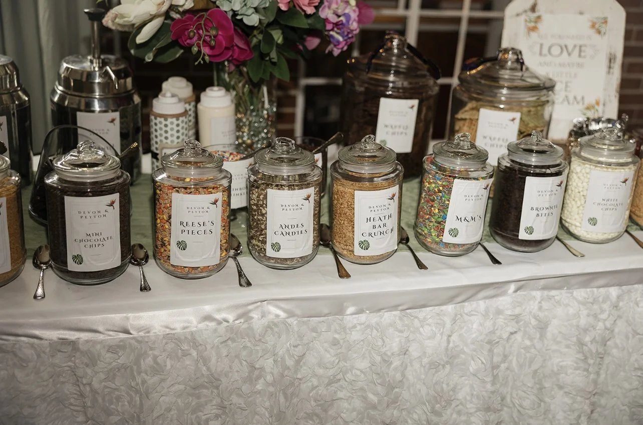 A table set with glass jars of various toppings, each labeled with different names such as 'Mini Chocolate Chips,' 'Reese's Pieces,' 'Andes Candies,' 'Health Bar Crunch,' 'M&M's,' 'Brownie Bites,' and 'White Chocolate Chips.' There is a bouquet of pi