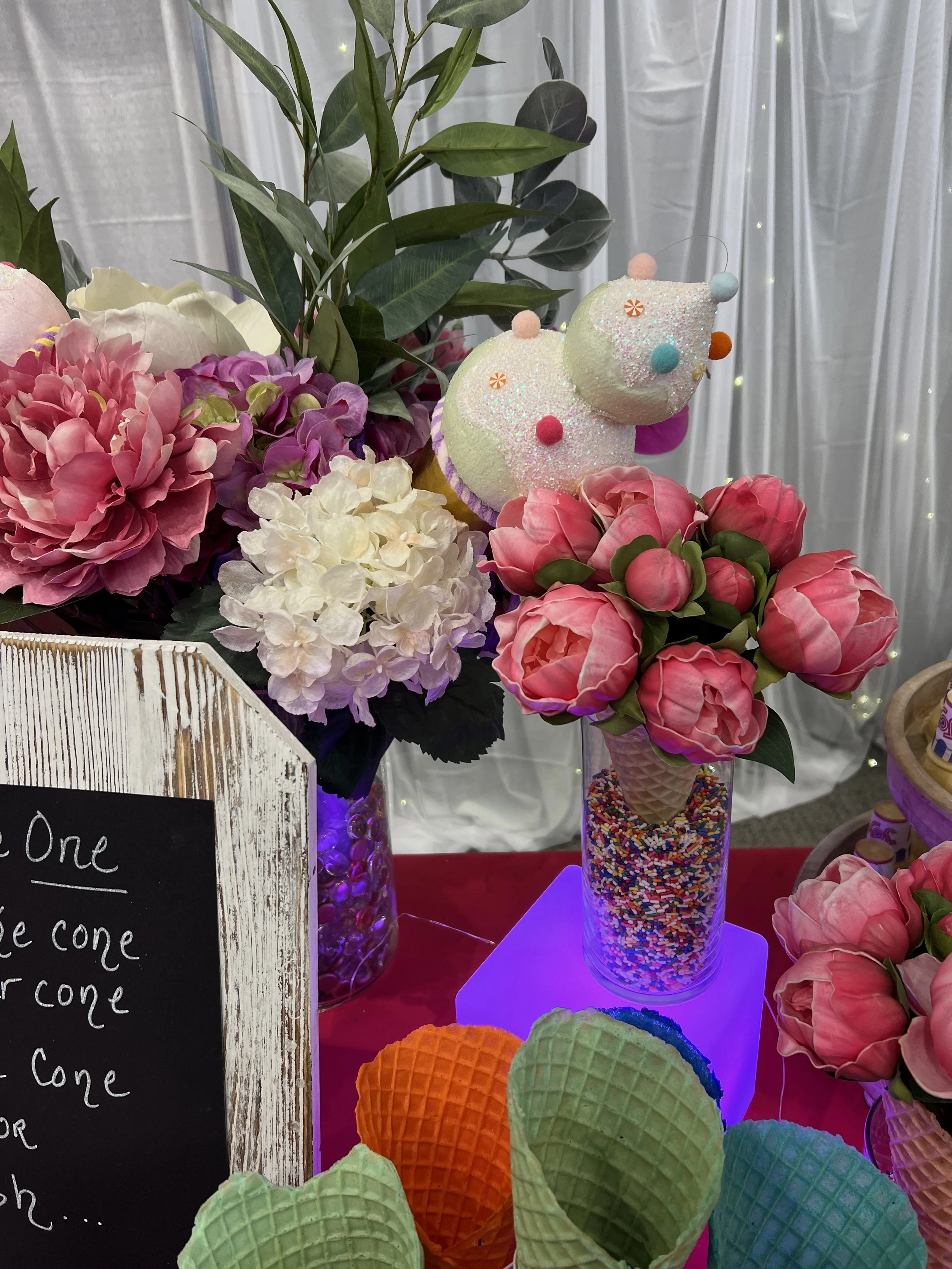 Colorful ice cream cones in orange, green, blue, and purple, displayed on a table with floral arrangements and decorative background lighting.