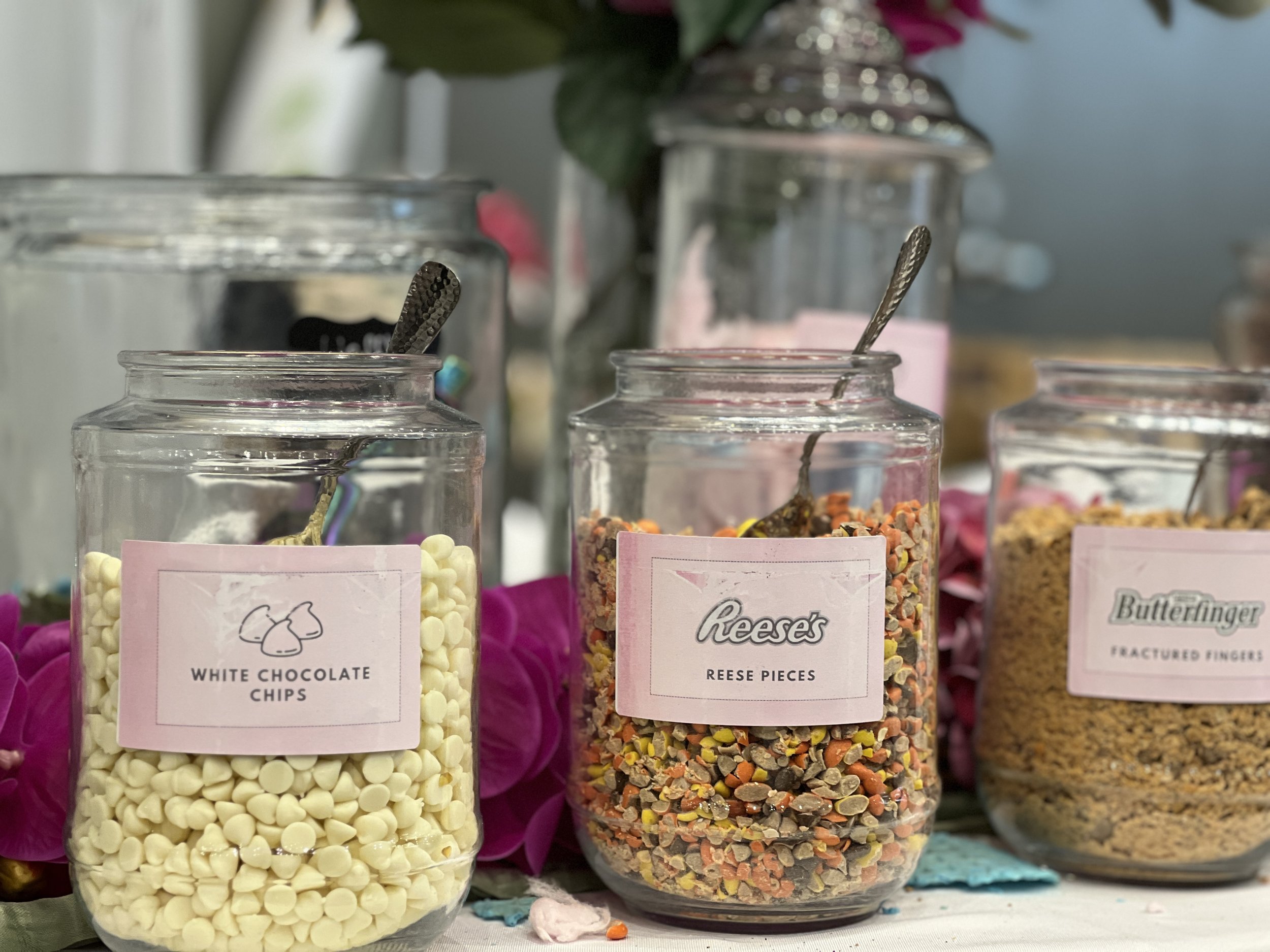 Three glass jars filled with candy toppings such as white chocolate chips, Reese's pieces, and crushed Fritos, with pink labels and spoons inside each jar.