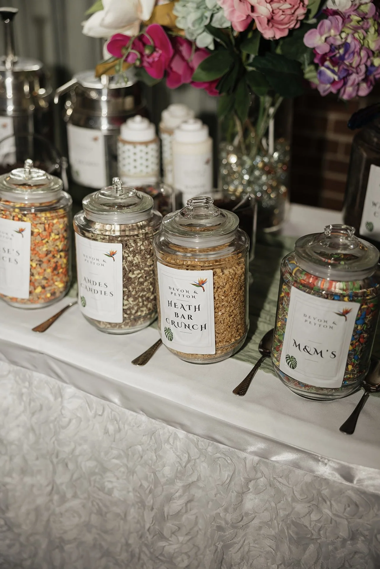 Jars of assorted candies and toppings on a white table, with a bouquet of pink, purple, and white flowers in the background.