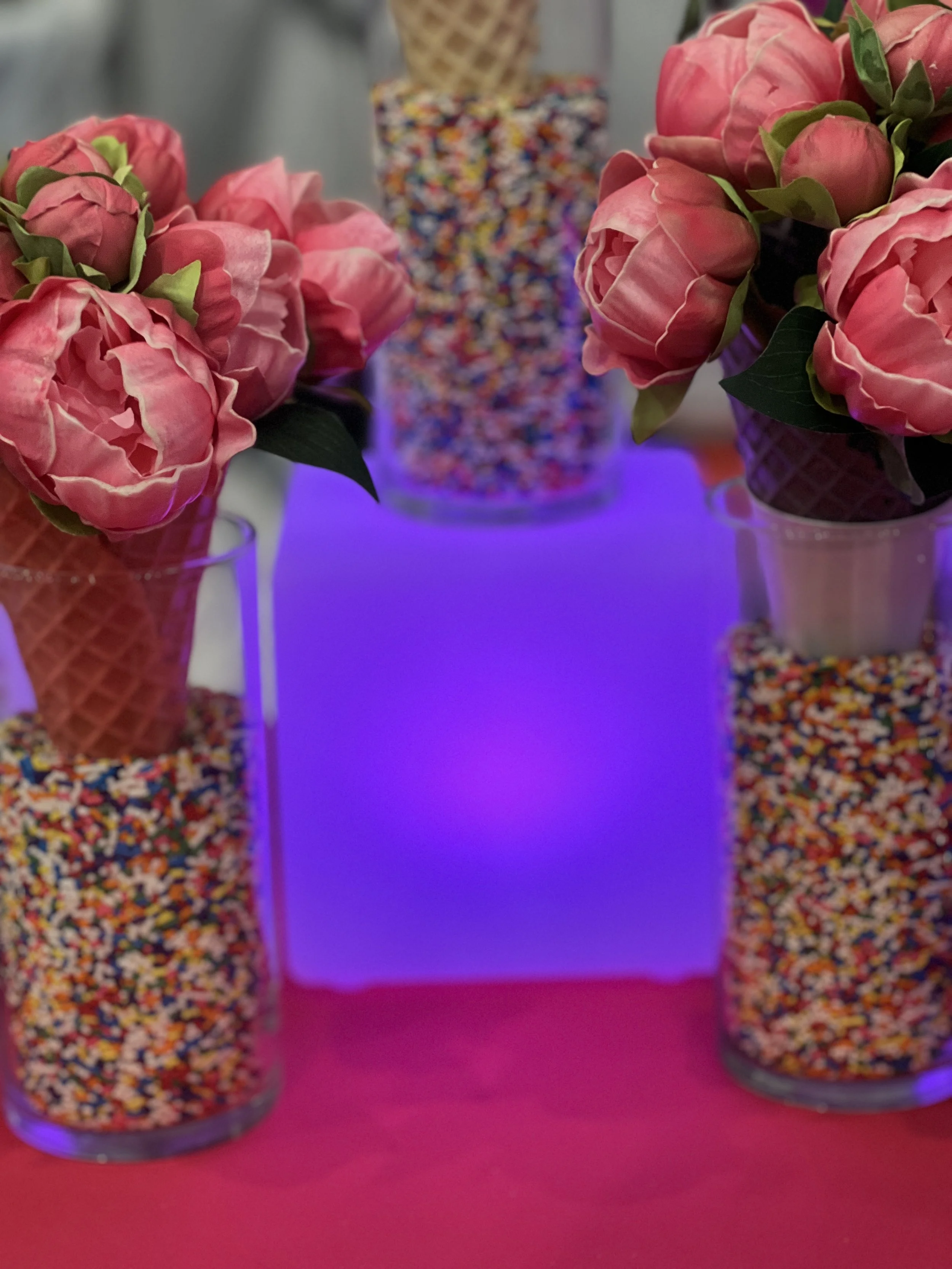 Three glass jars filled with colorful sprinkles on a pink surface, with two pink flowers on either side and a purple-lit background.