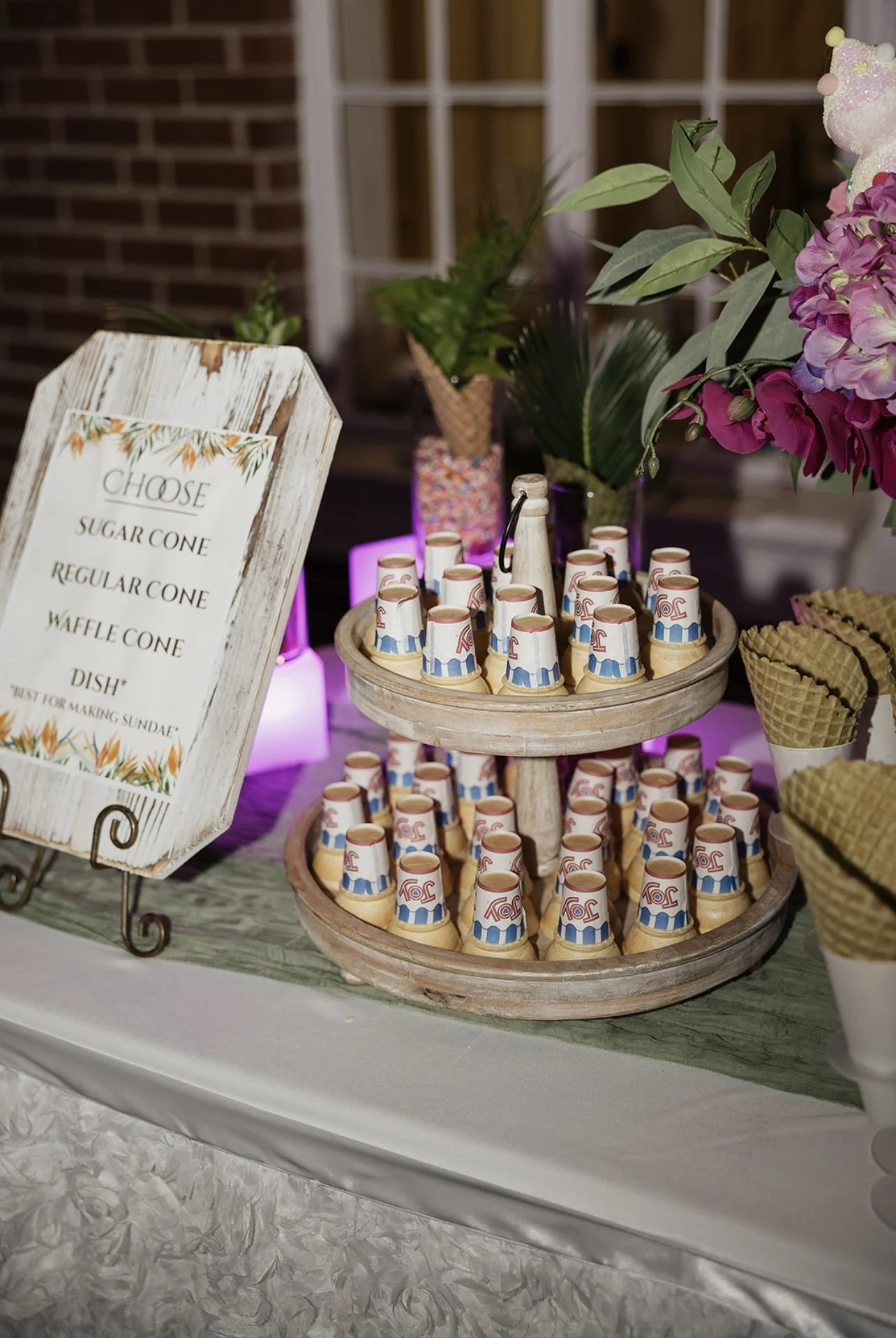 A dessert table with a wooden stand holding chocolate cups decorated with a red, white, and blue flag pattern, surrounded by a floral arrangement with pink and purple flowers, and a sign listing different cone options like sugar cone, regular cone, a