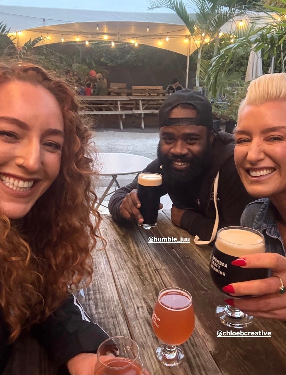 Three friends happily enjoying drinks at an outdoor bar or restaurant during the evening. Two women and one man are smiling, each holding glasses of dark beer and a pinkish cocktail, with string lights overhead and greenery around.