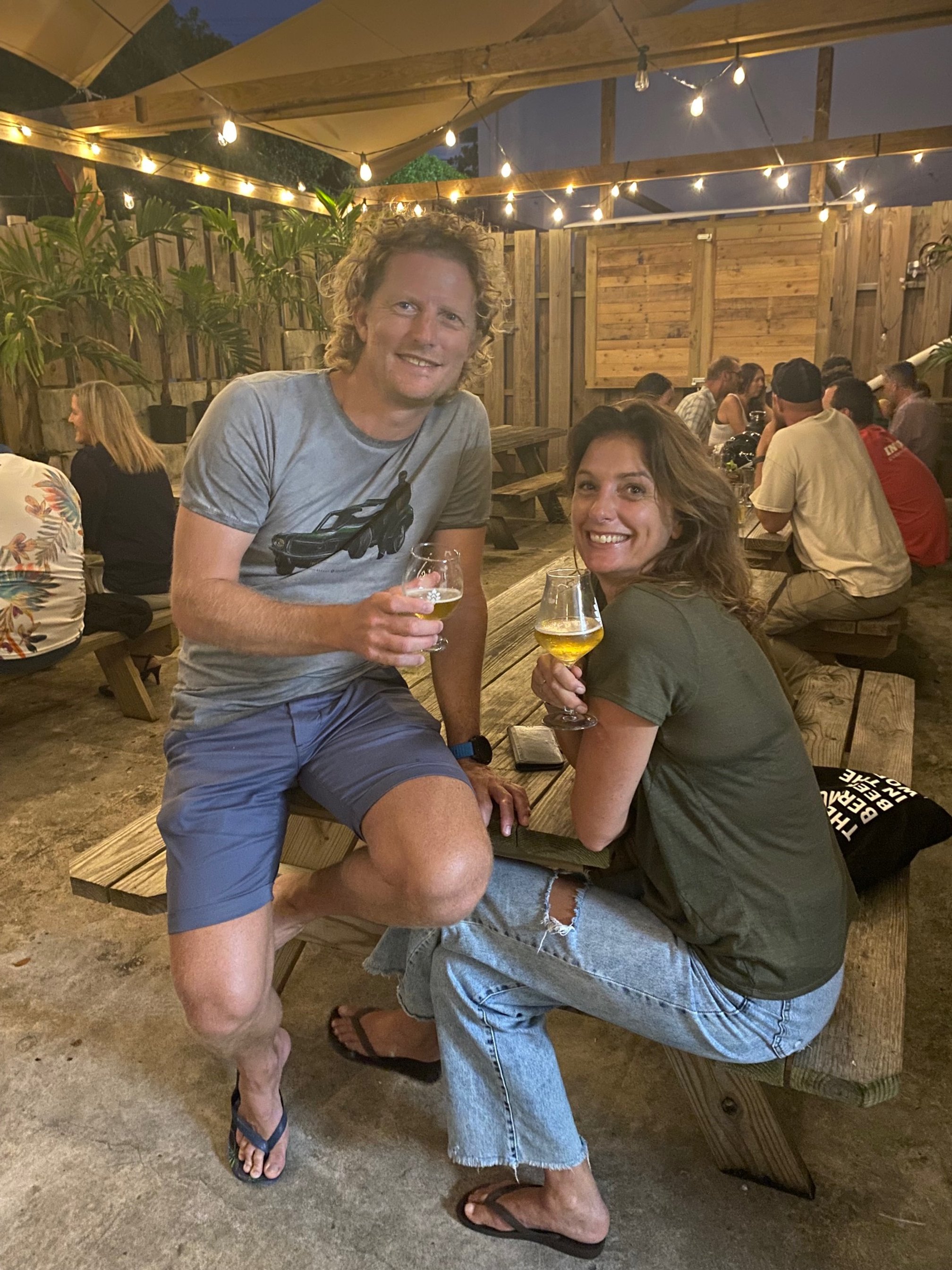 Two friends are sitting at a wooden picnic table outdoors under string lights, smiling and holding drinks. The man is standing with one leg on the bench, wearing a gray t-shirt, gray shorts, and flip-flops. The woman is sitting with her knees up, wearing a green t-shirt, ripped jeans, and sandals. In the background are other people sitting at picnic tables, a wooden fence, and plants.
