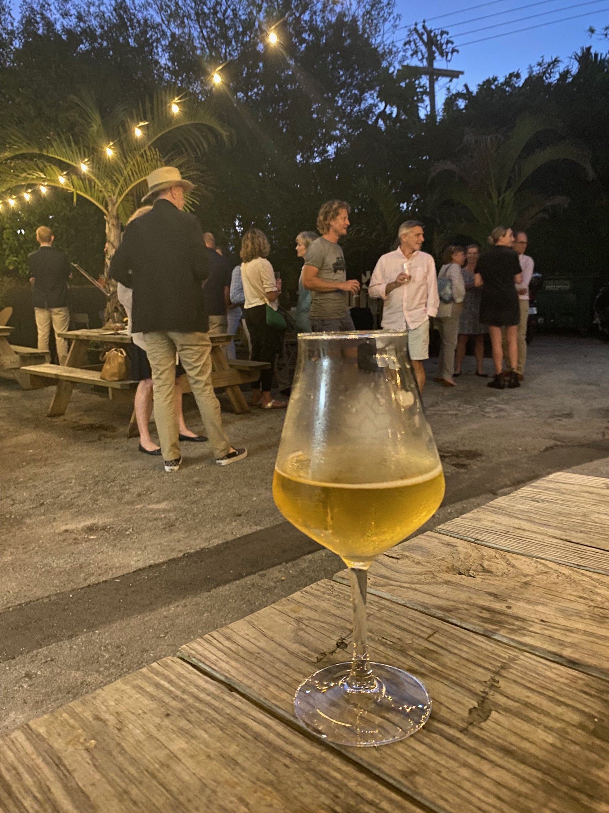 A glass of white wine or champagne on a wooden table with a group of people socializing outdoors during the evening. The scene is illuminated by string lights, with trees and power lines visible in the background.